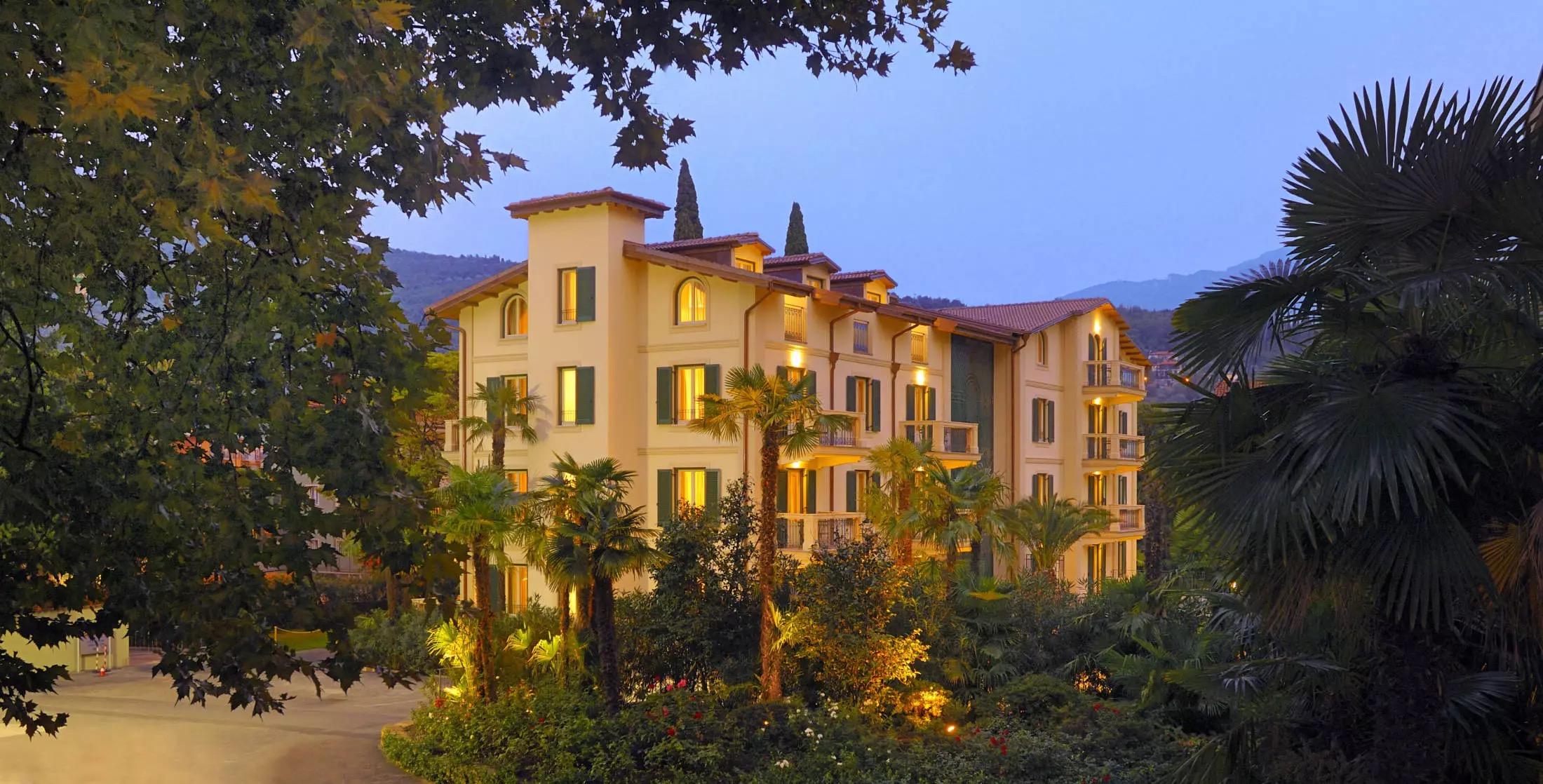 The front of a cream-coloured hotel building at dusk, with its windows lit, green shutters, a terracotta roof, and palm trees in front.