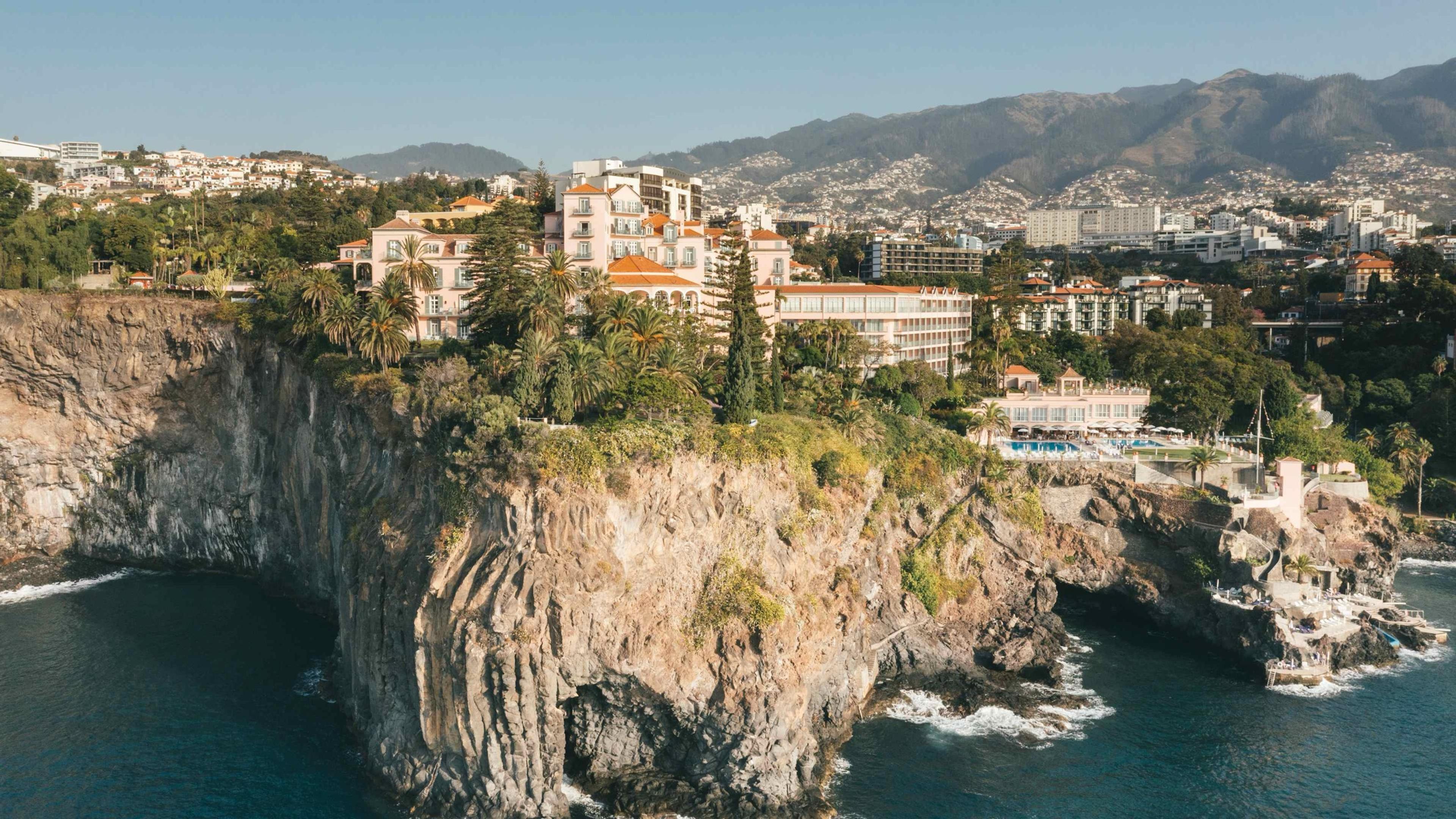 An aerial view shows the hotel's pink buildings and gardens on a cliff above the sea, a pool terrace at its foot, with Funchal and the mountains behind.