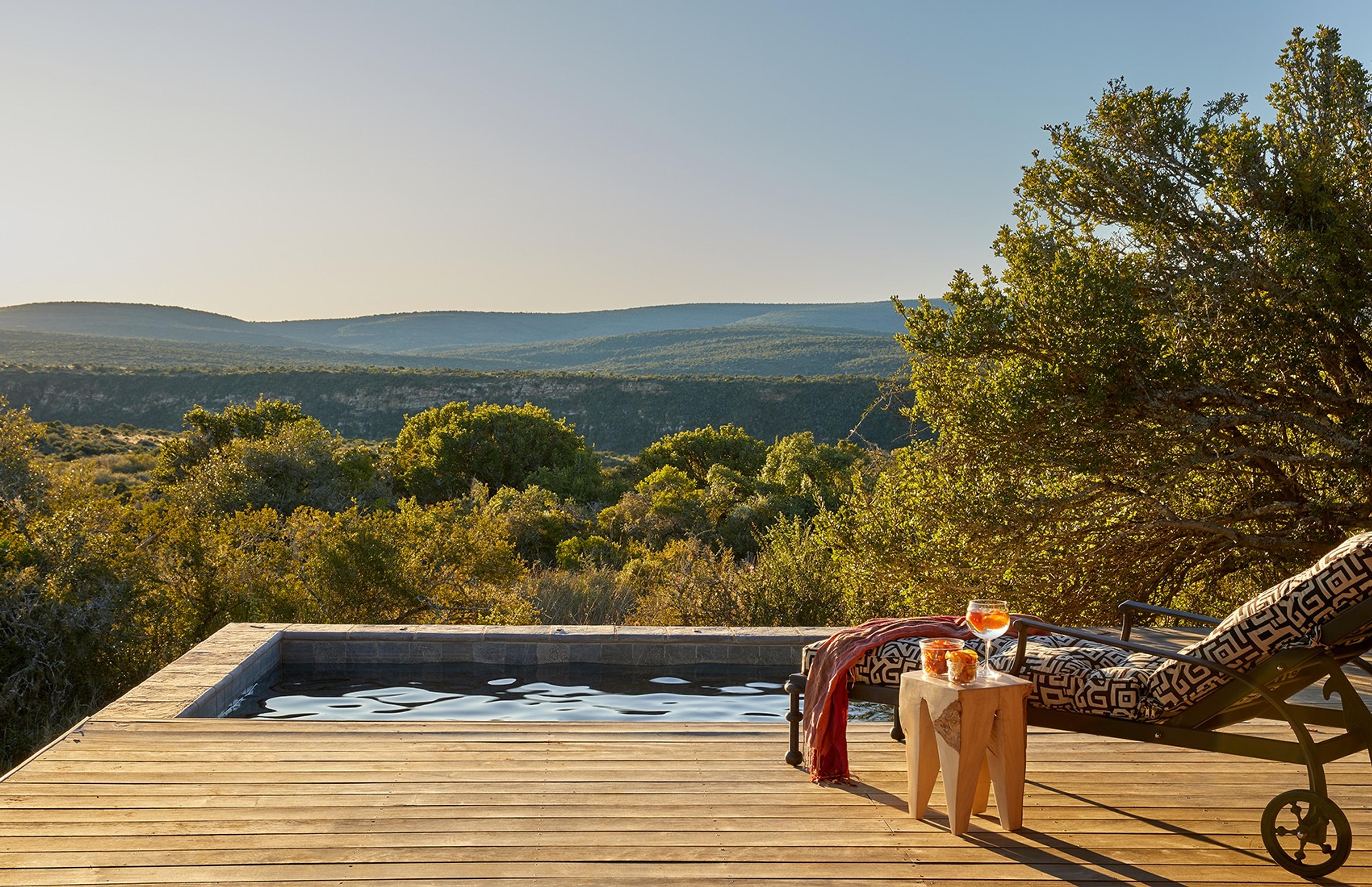 A small plunge pool on a wooden deck overlooking scrubland and distant hills, with a sun lounger and a drink on a side table.