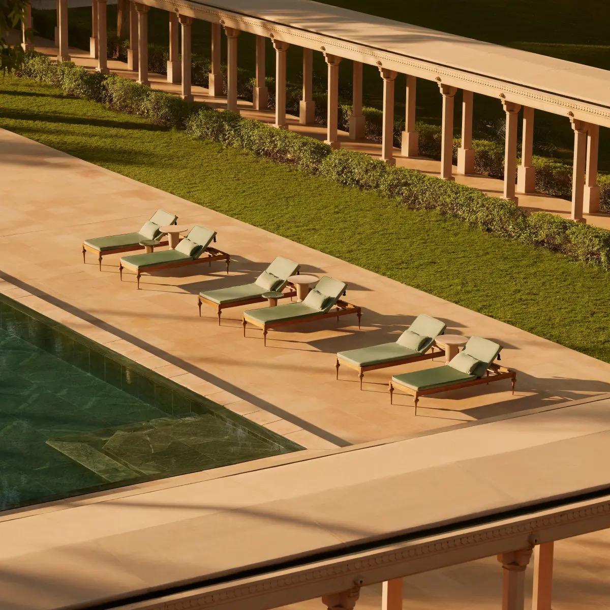 An aerial view of a dark tiled pool beside a row of green sun loungers on a stone terrace, with a colonnade and lawn above.