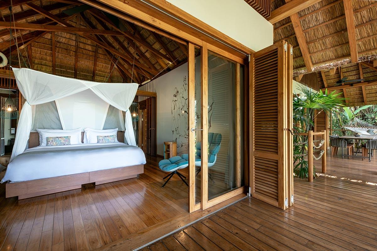 a canopied bed with white linen in a thatched-roof bedroom, its wooden shutters open to a private deck with a hammock among trees