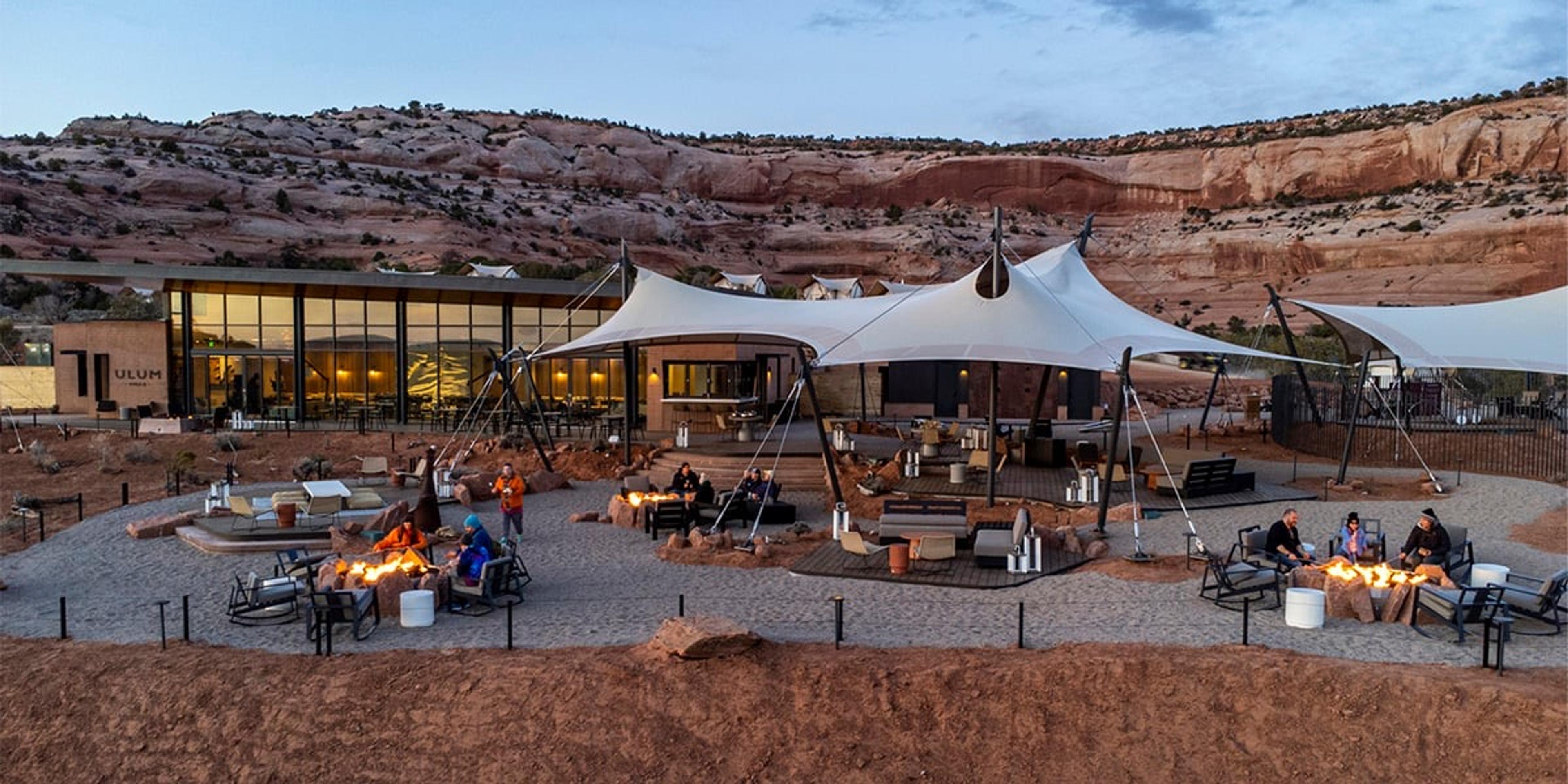 A long wooden dining table set with bowls, glasses and cutlery, with pendant lamps overhead and floor-to-ceiling windows showing desert and a rock arch in the distance. A lounge area is at the far end.