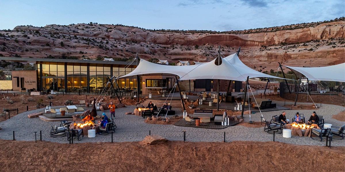 A long wooden dining table set with bowls, glasses and cutlery, with pendant lamps overhead and floor-to-ceiling windows showing desert and a rock arch in the distance. A lounge area is at the far end.