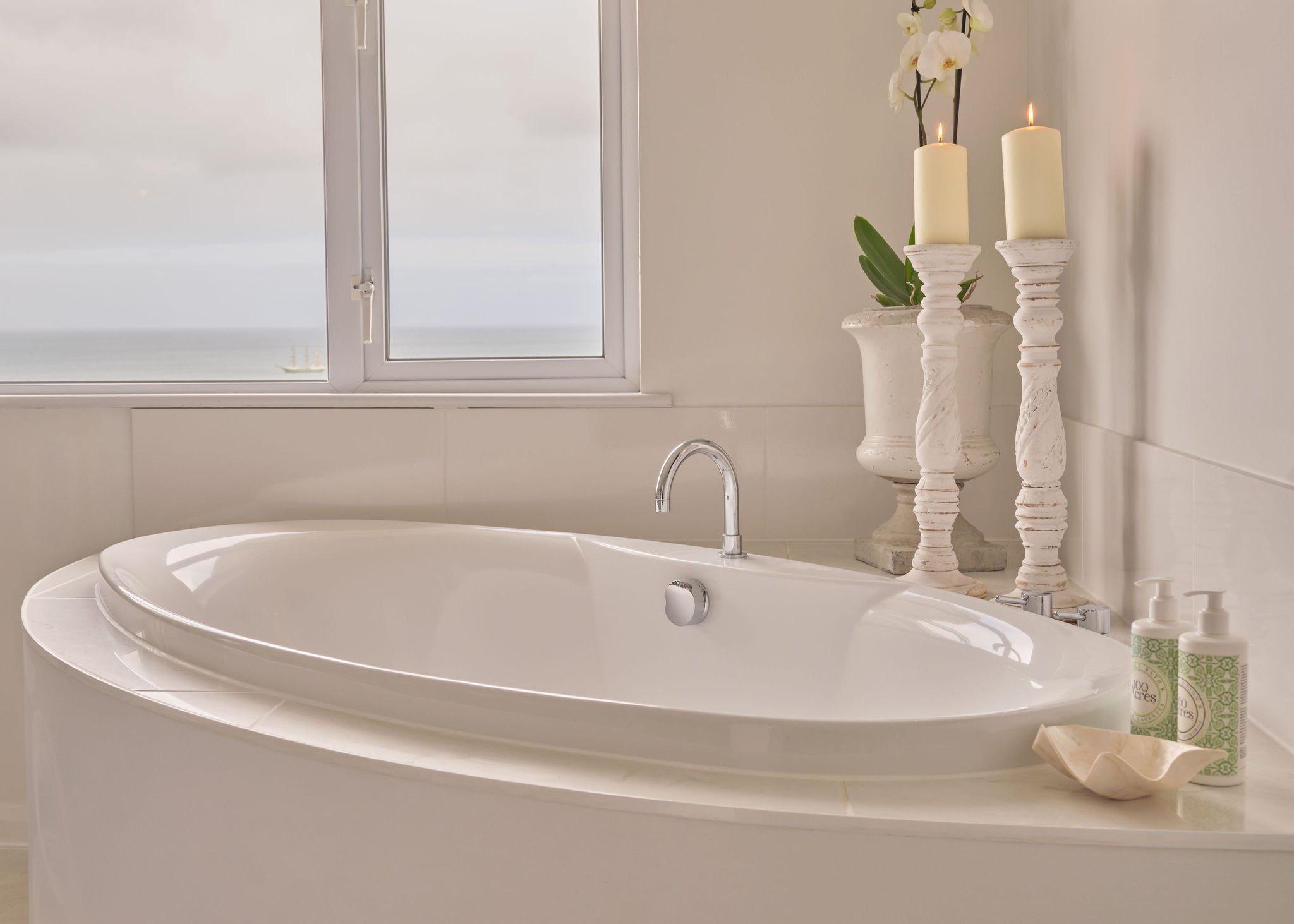 A freestanding bathtub beside a window with the sea and a sailing boat visible, two lit candles and a white orchid on the surround.