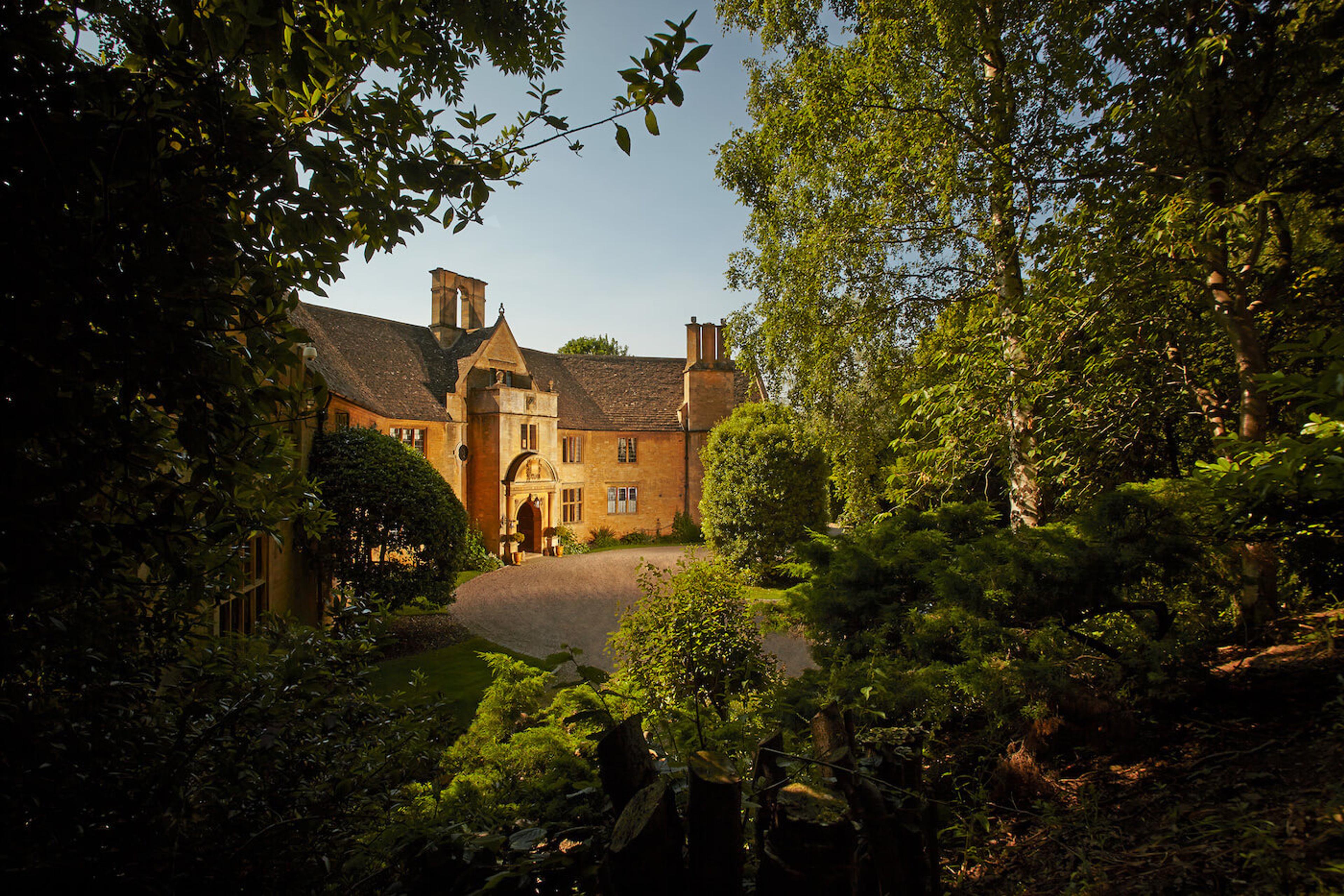 A honey-stone manor house seen through trees in late afternoon light, with an archway entrance and a gravel forecourt.