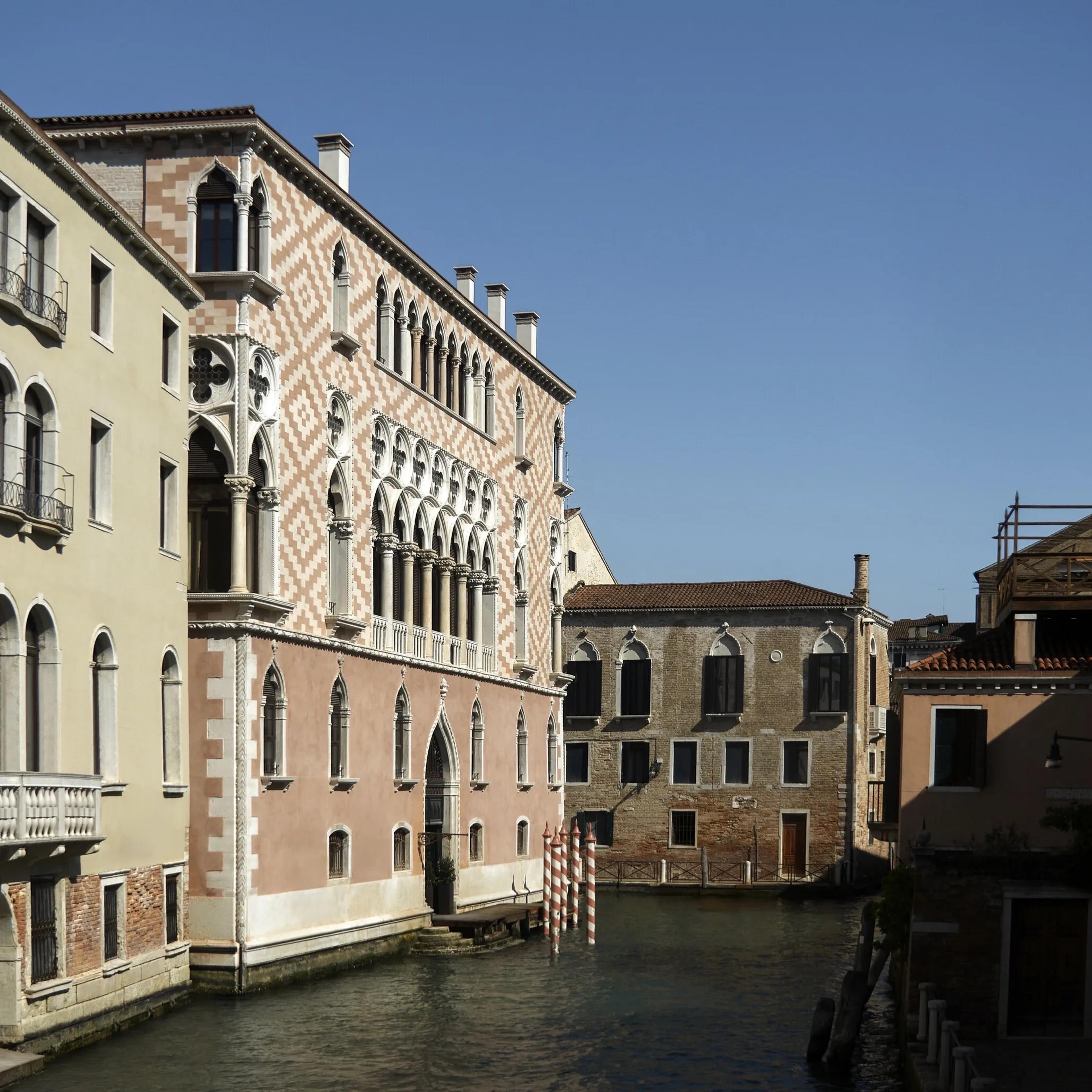 A pink and white Gothic palazzo facade rising from a Venice canal, with striped mooring poles in front and plainer buildings alongside