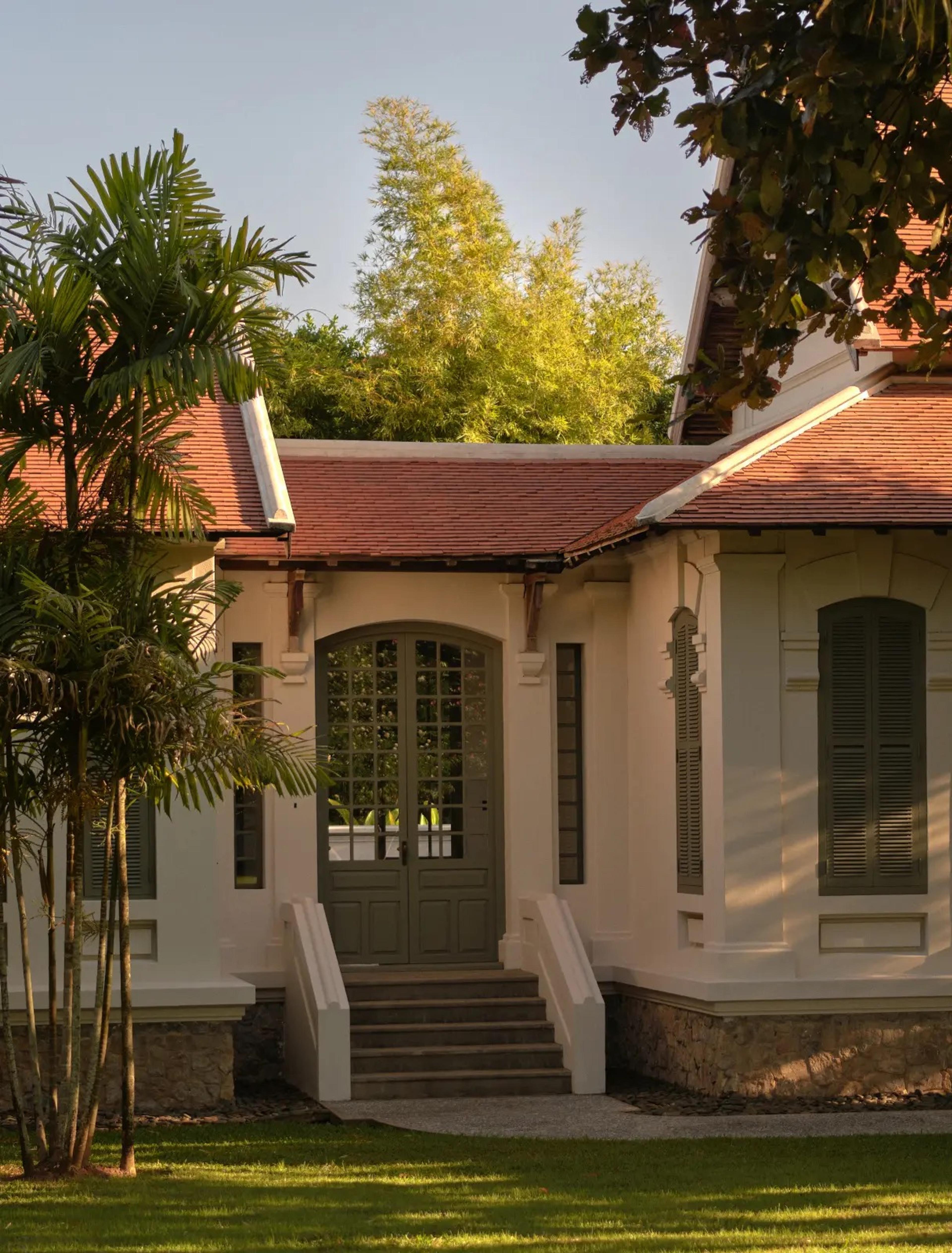 A white building with a red tiled roof and a columned entrance, a short flight of steps leading to double doors, framed by a palm on one side.