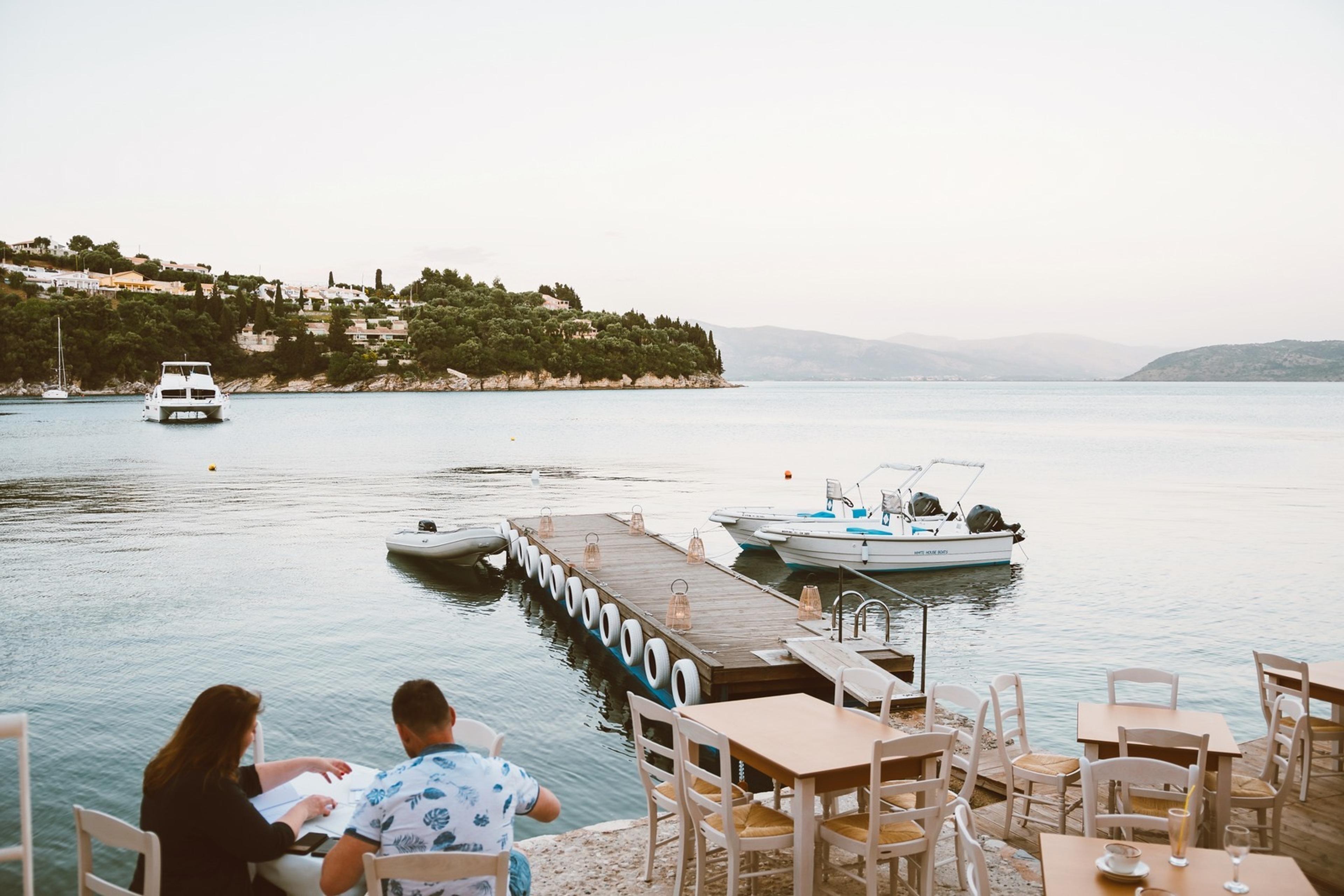 Diners at waterside tables beside a timber jetty on Kalami Bay, with small boats moored and wooded hills across the water