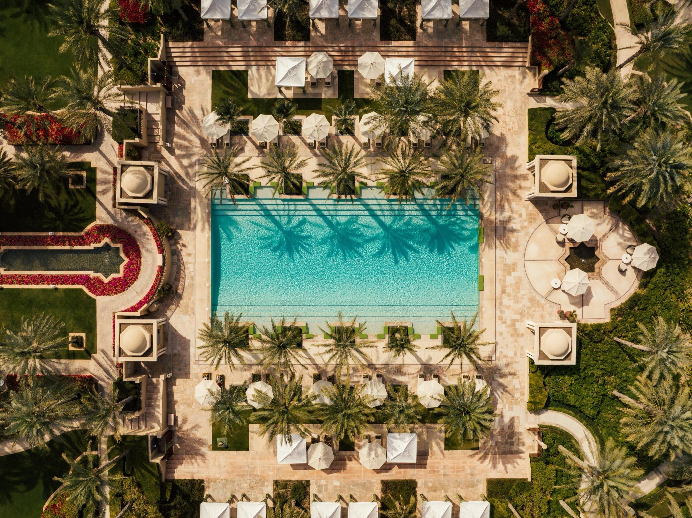 An aerial view straight down on a rectangular pool shaded by rows of palm trees, flanked by domed cabanas and gardens.