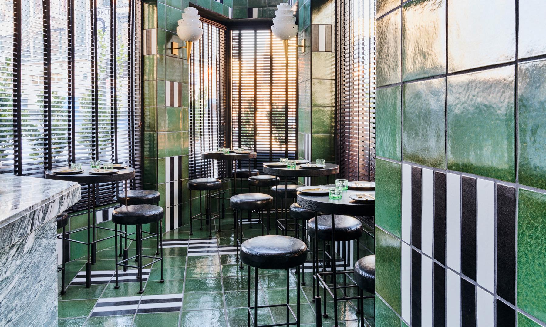 A narrow green-tiled bar nook with round black stools, striped wood blinds and a grey marble counter in the foreground.