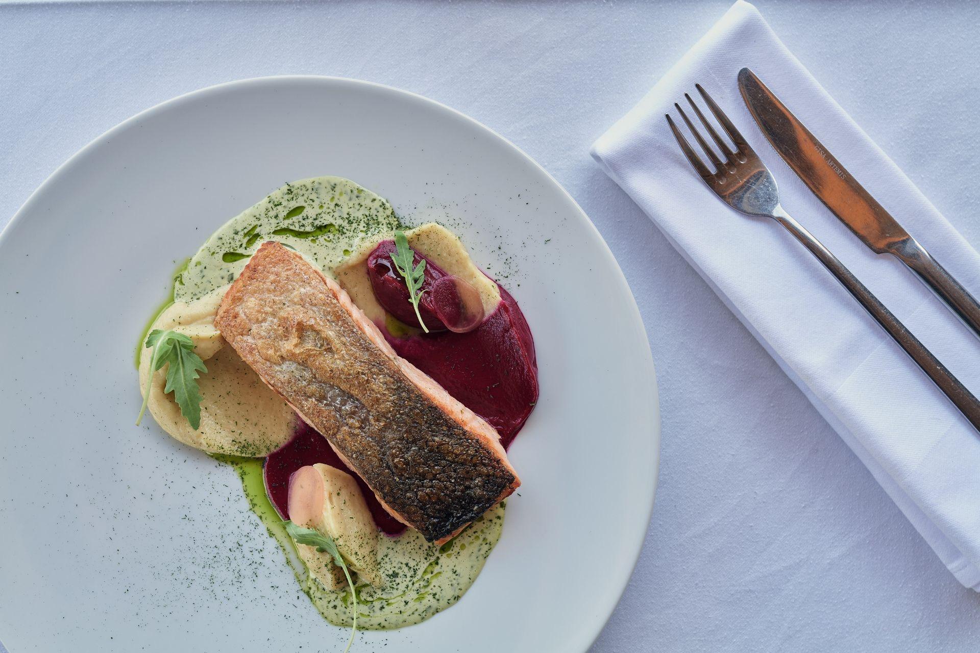 A plated fillet of fish with pureed accompaniments, beside a folded napkin and cutlery on a white tablecloth.