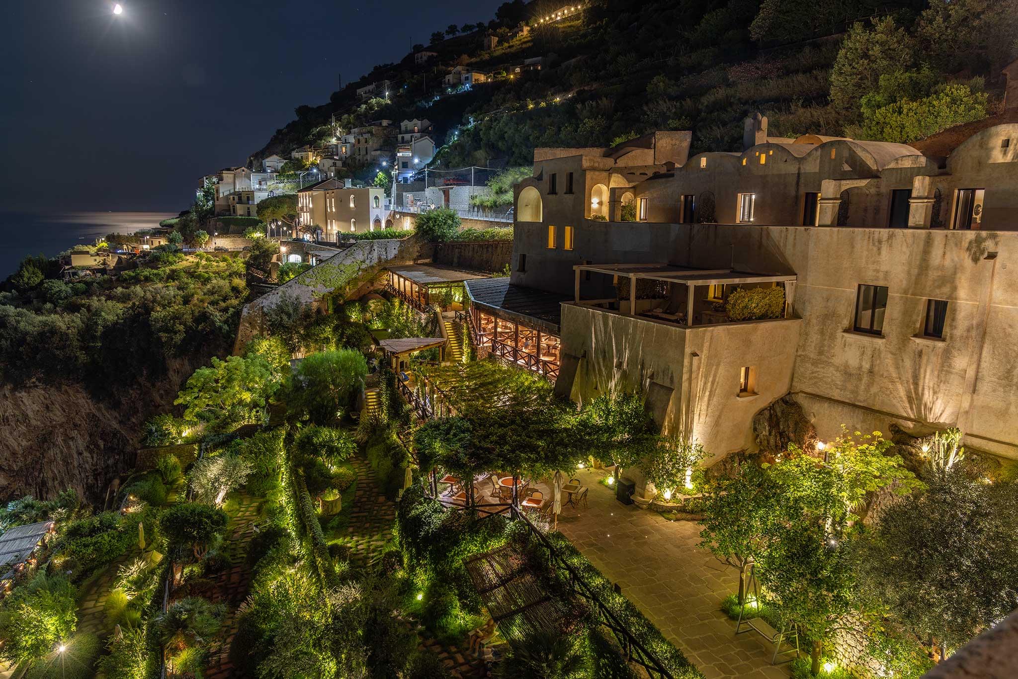 an aerial night view of the hotel built into a hillside above the coast, its windows lit, with a lit coastal village beyond and the moon overhead