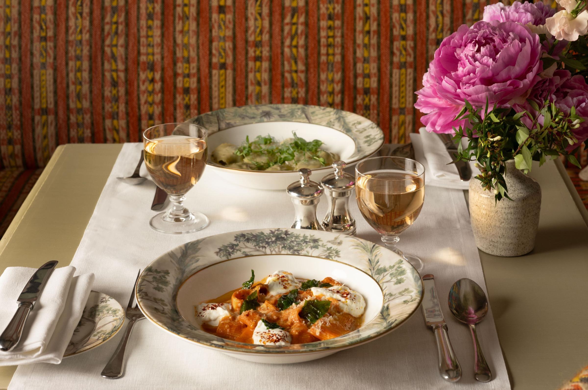Two plated dishes and glasses of white wine on a table laid with a patterned cloth, with a vase of pink peonies beside them.