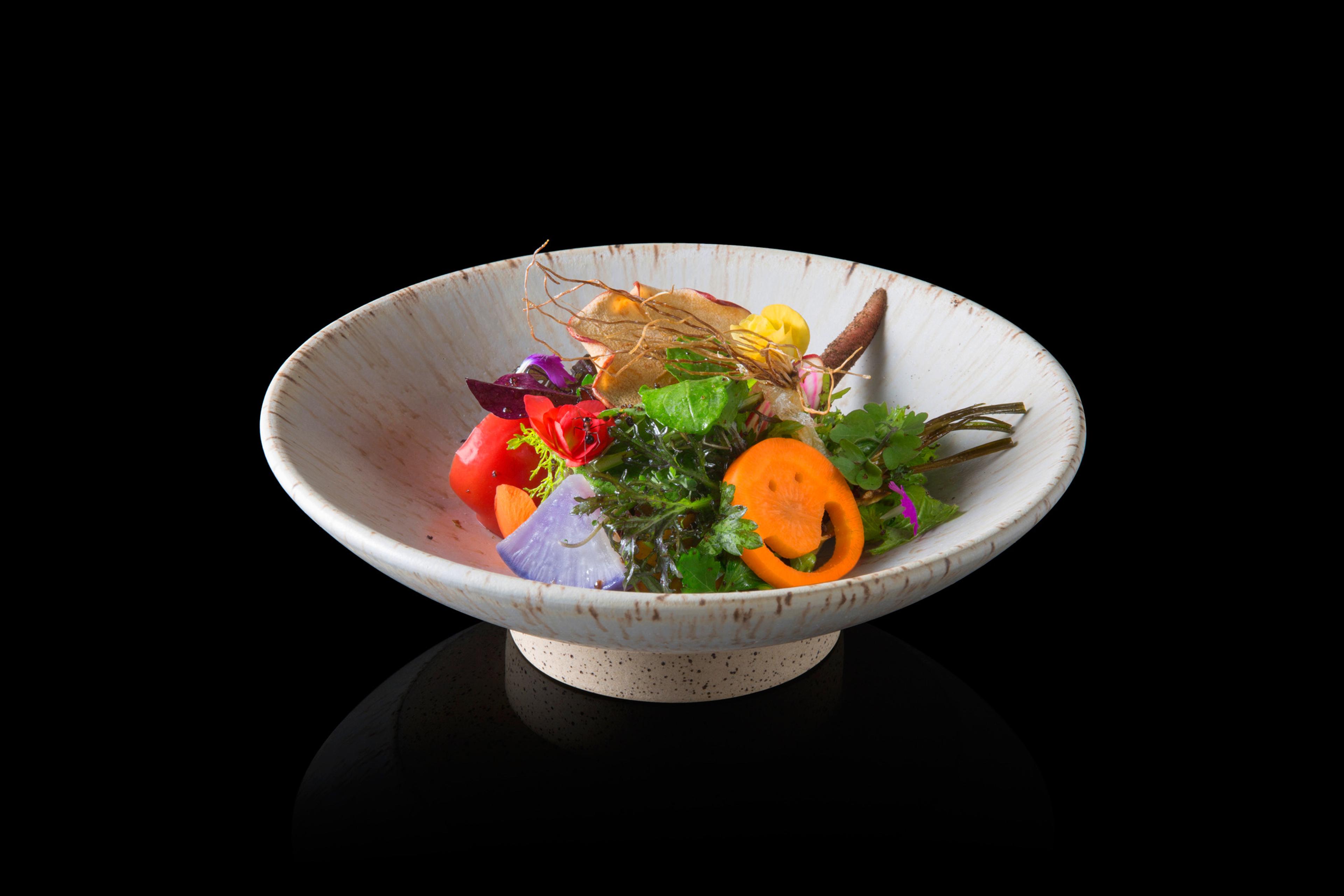 A shallow bowl of salad leaves, carrot and flowers against a black background