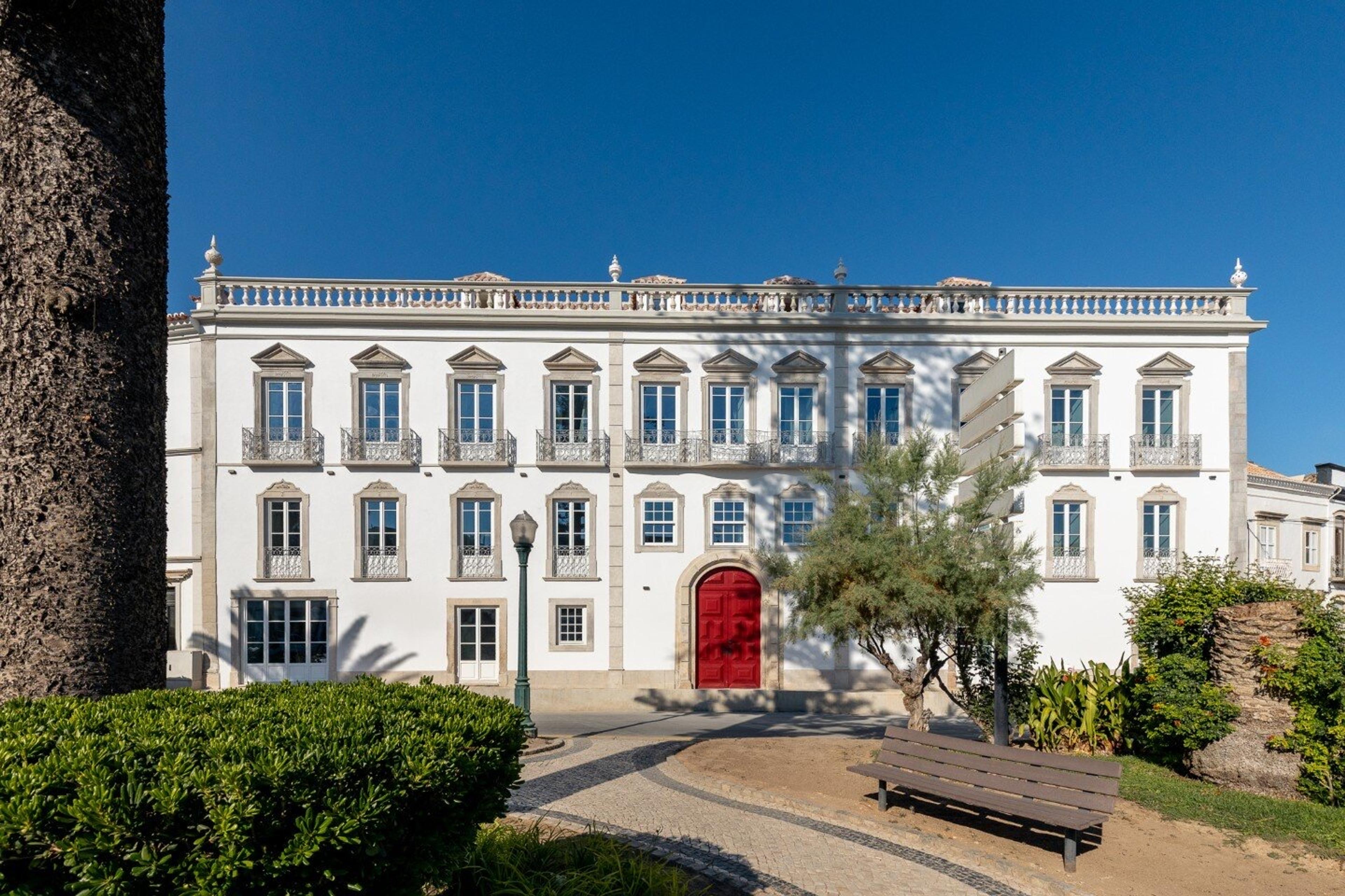 A white three-storey palace facade with balconied windows and a stone balustrade along the roofline, a red double door at its centre, seen across a garden with a bench and trees.