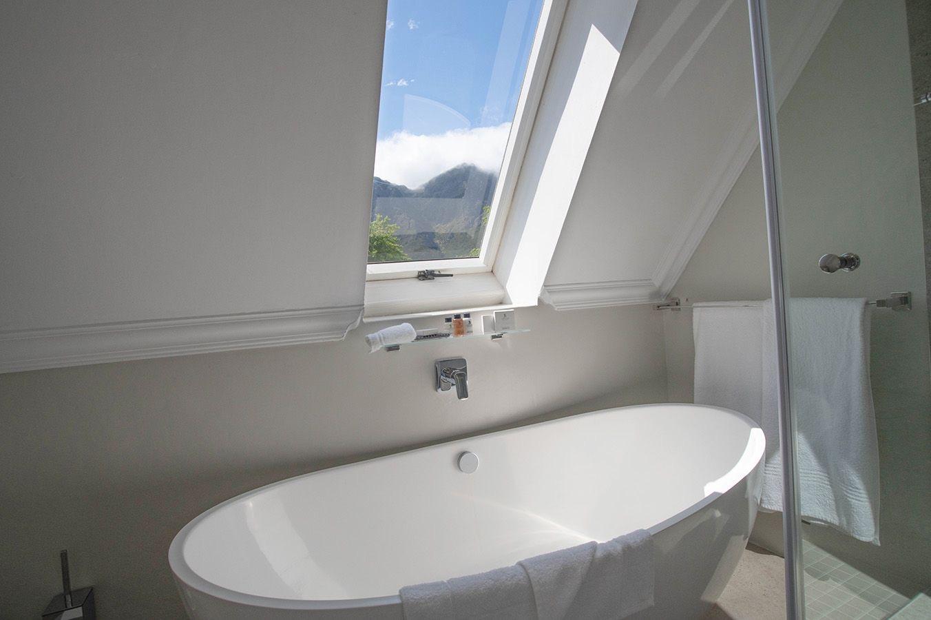 A freestanding white bath under a roof window, with a mountain visible through the glass.