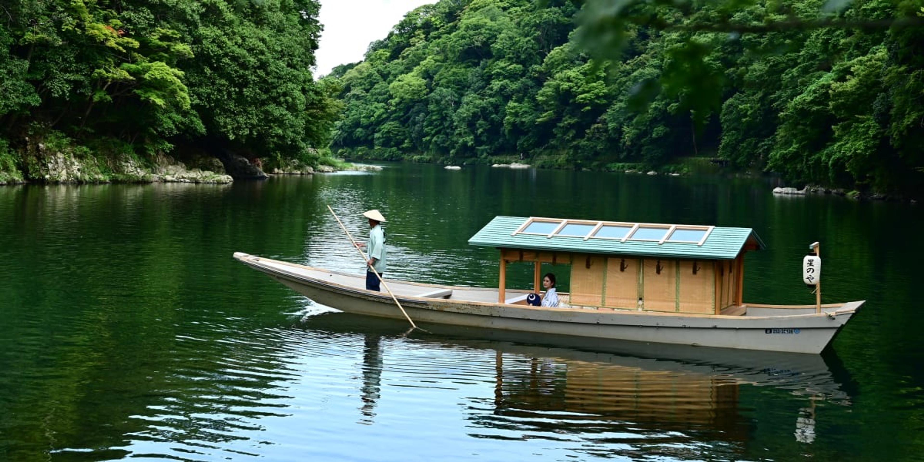 A covered wooden boat on the river below Arashiyama's wooded hills, a boatman standing at the stern