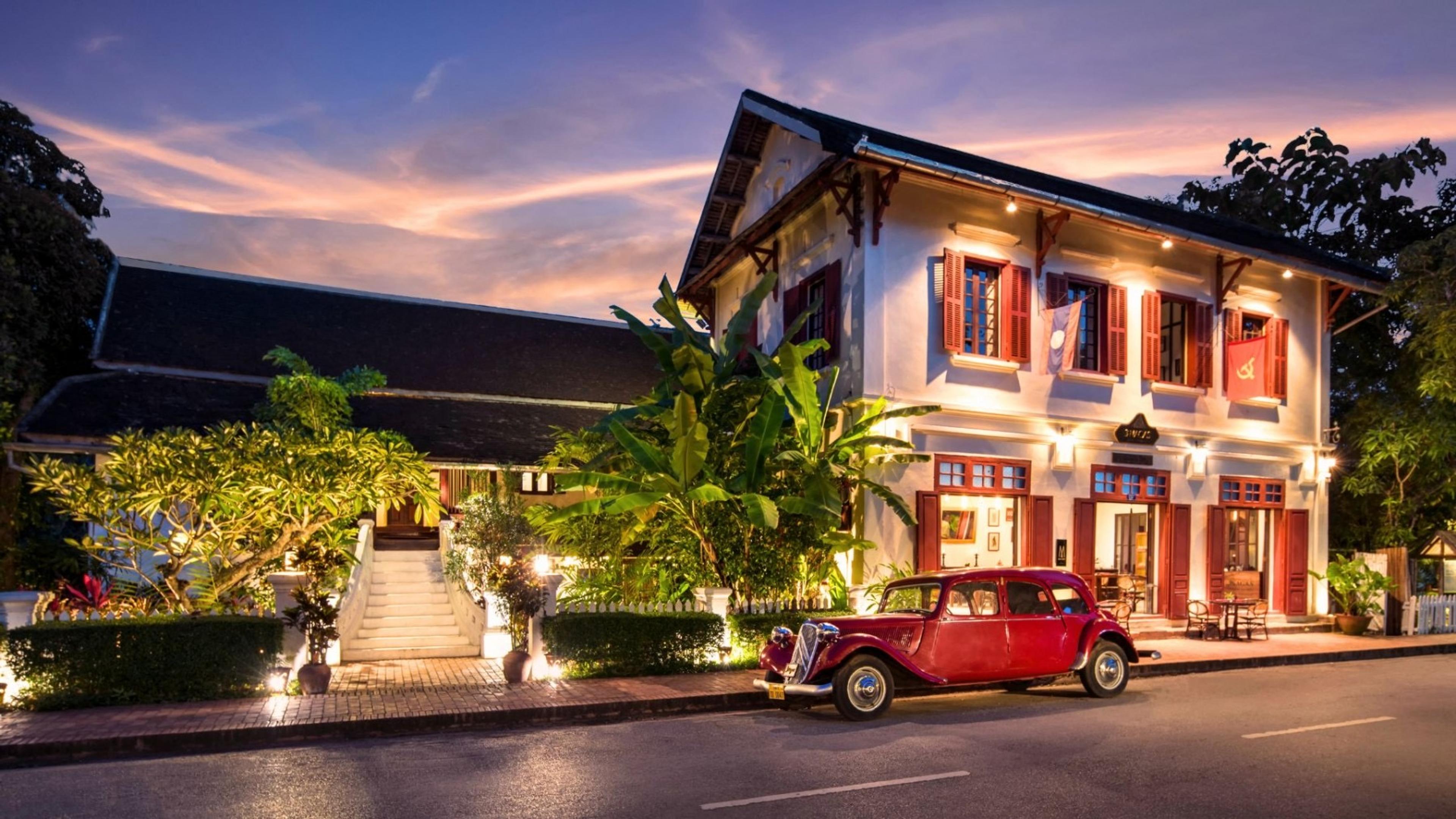 A two-storey colonial building lit up at dusk, with a vintage red car parked on the street in front and palm leaves framing the scene.