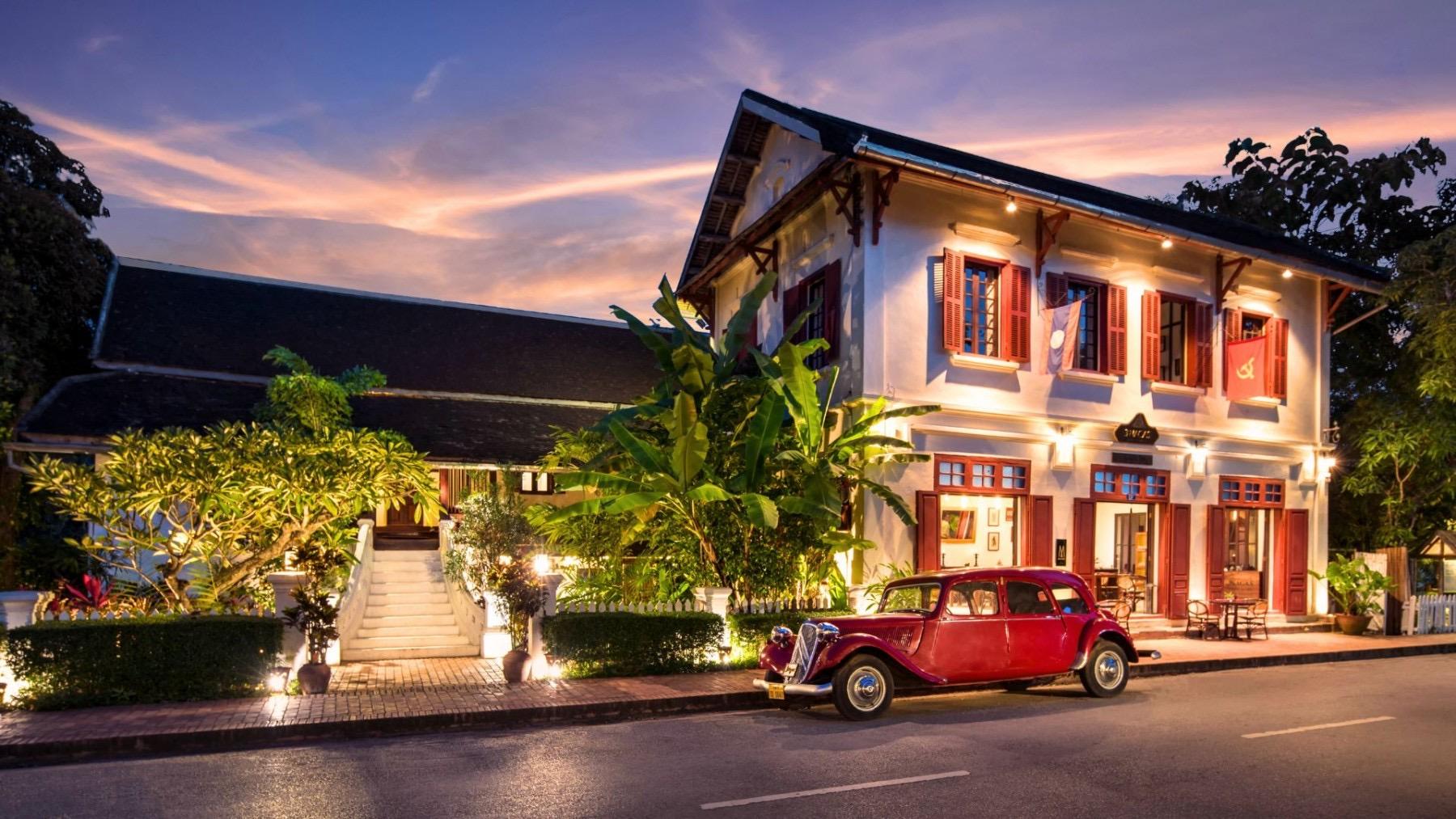 A two-storey colonial building lit up at dusk, with a vintage red car parked on the street in front and palm leaves framing the scene.