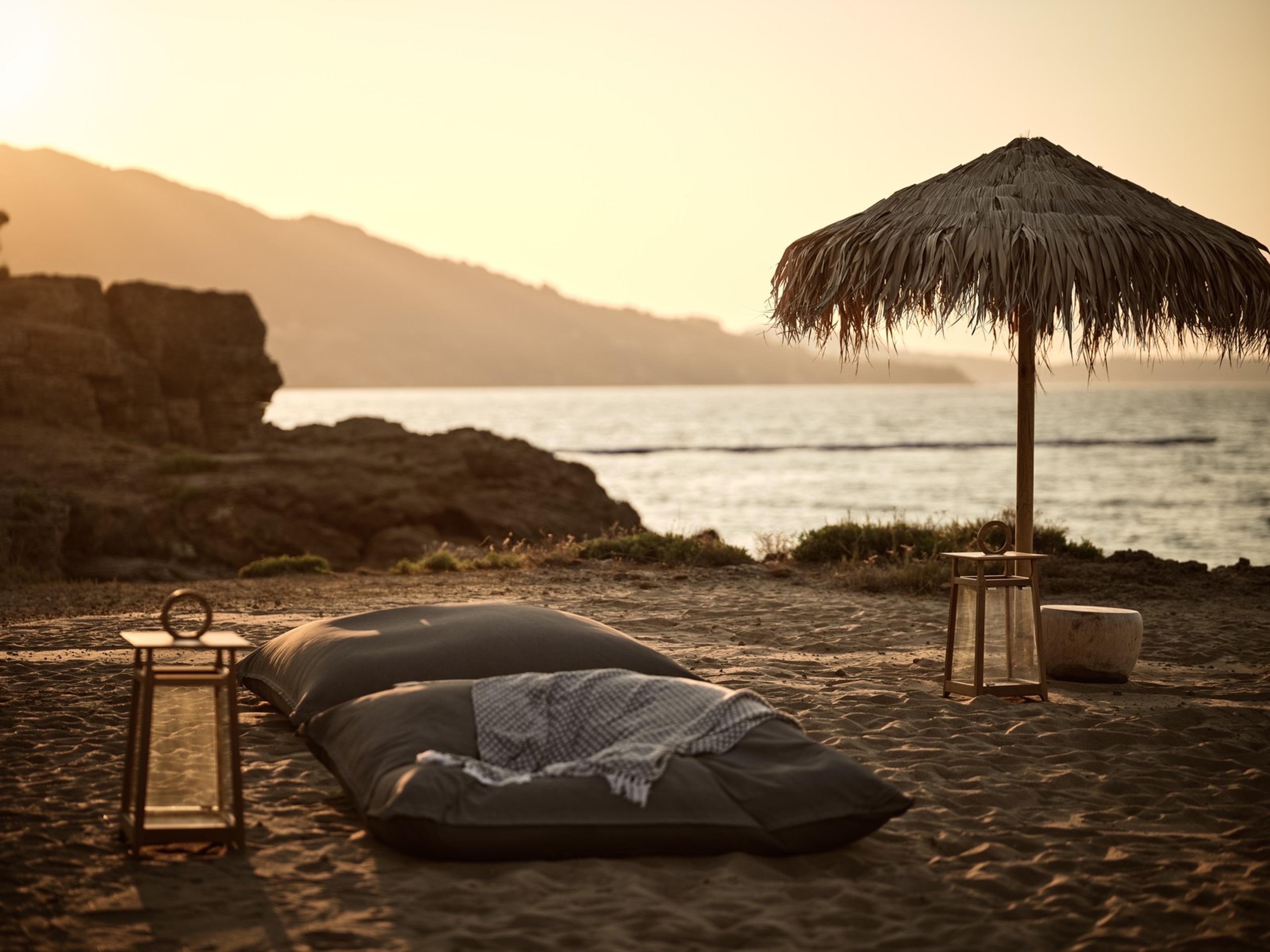 Floor cushions and a lantern on a beach at dusk, beside a thatched umbrella and rocks above the sea.