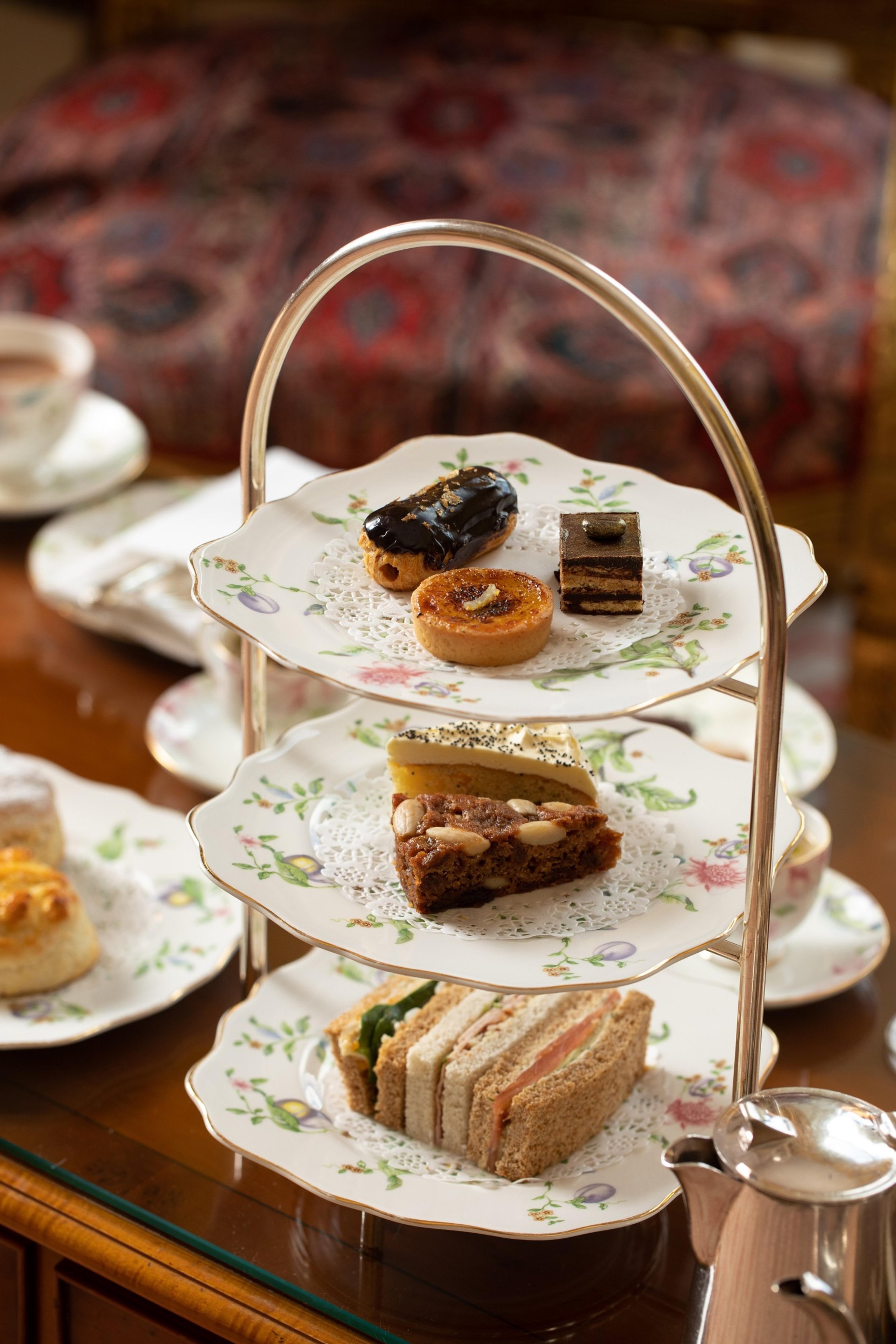 A three-tier stand of afternoon tea cakes, tarts and sandwiches on patterned china, with a teapot and cup nearby.