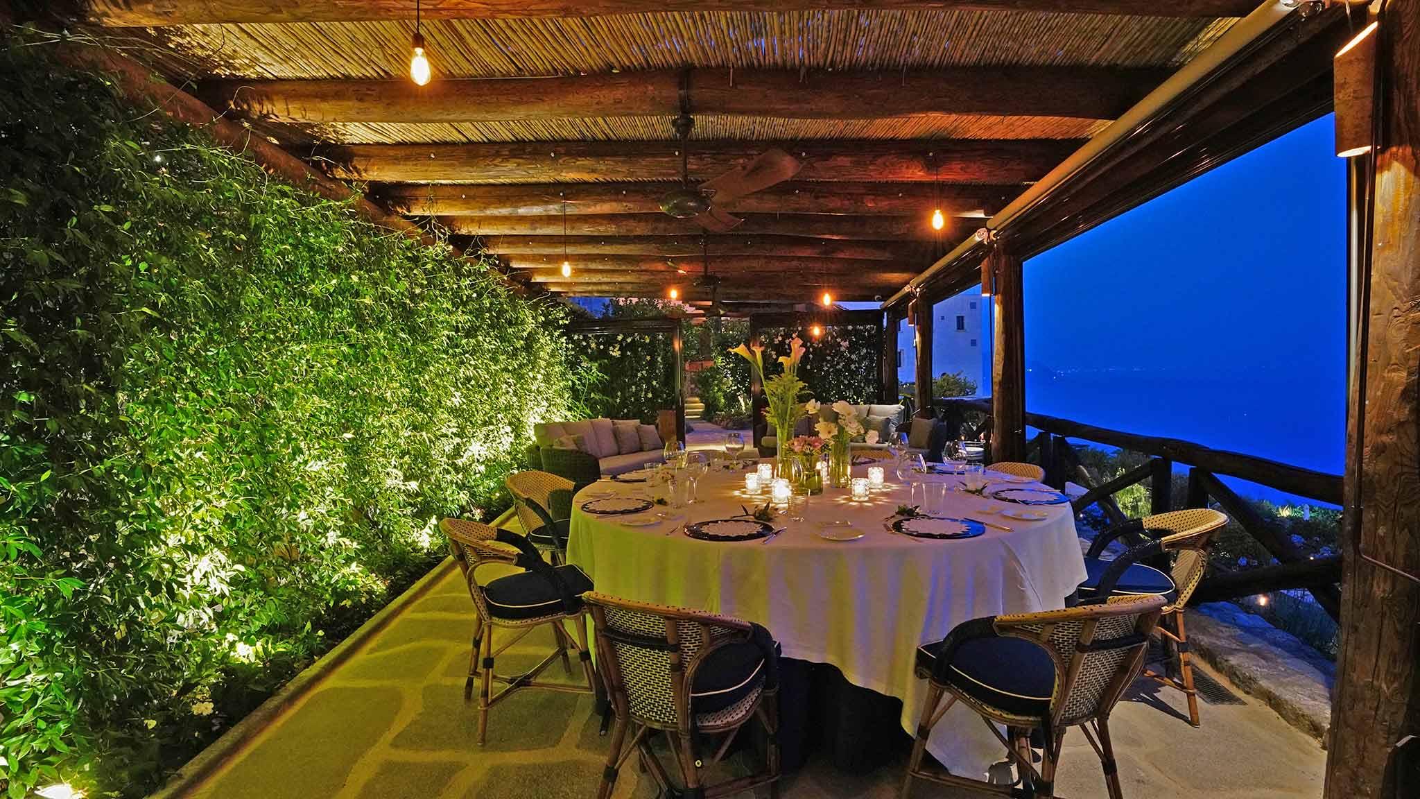 a round table laid for dinner on a covered terrace at dusk, strung lights above, a wall of climbing greenery to one side and the sea visible through the open side