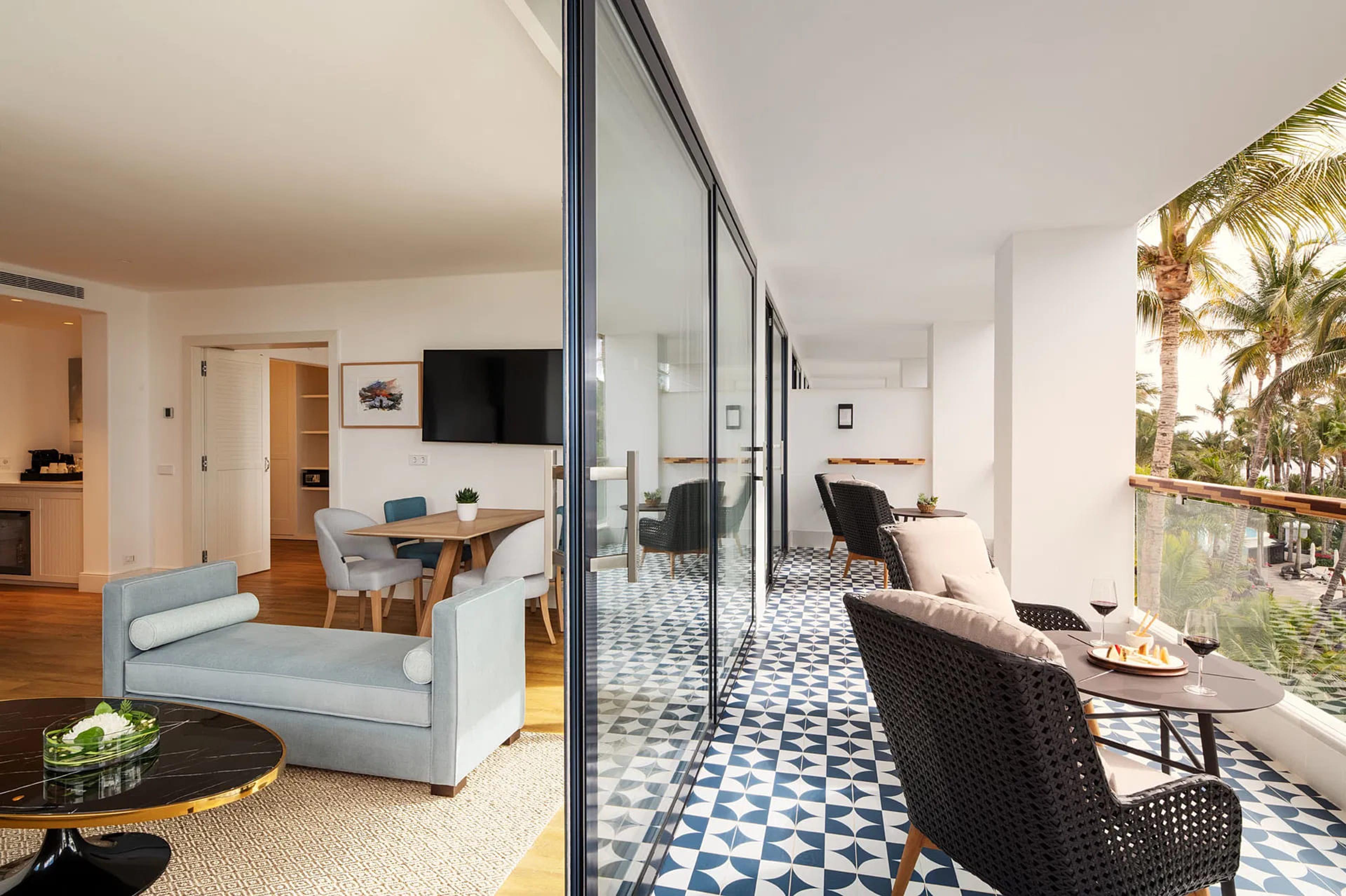 A hotel suite's living area with a pale sofa and dining table opens onto a tiled balcony set with wine glasses and a plate of food, palm trees beyond the railing.