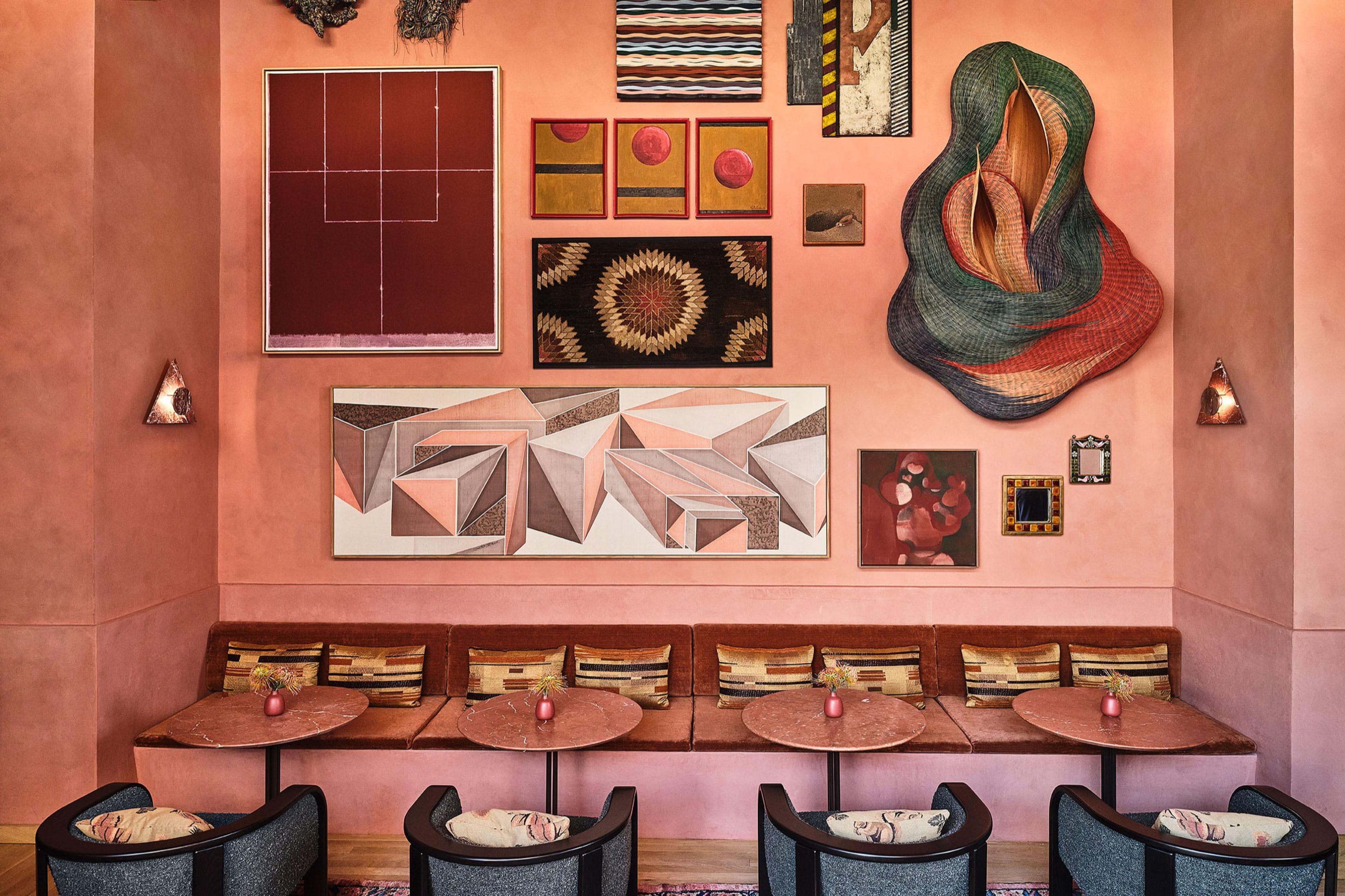 A pink plastered lounge with a built-in banquette, round marble-topped tables, and a wall hung with a dozen small abstract artworks and woven textile pieces.