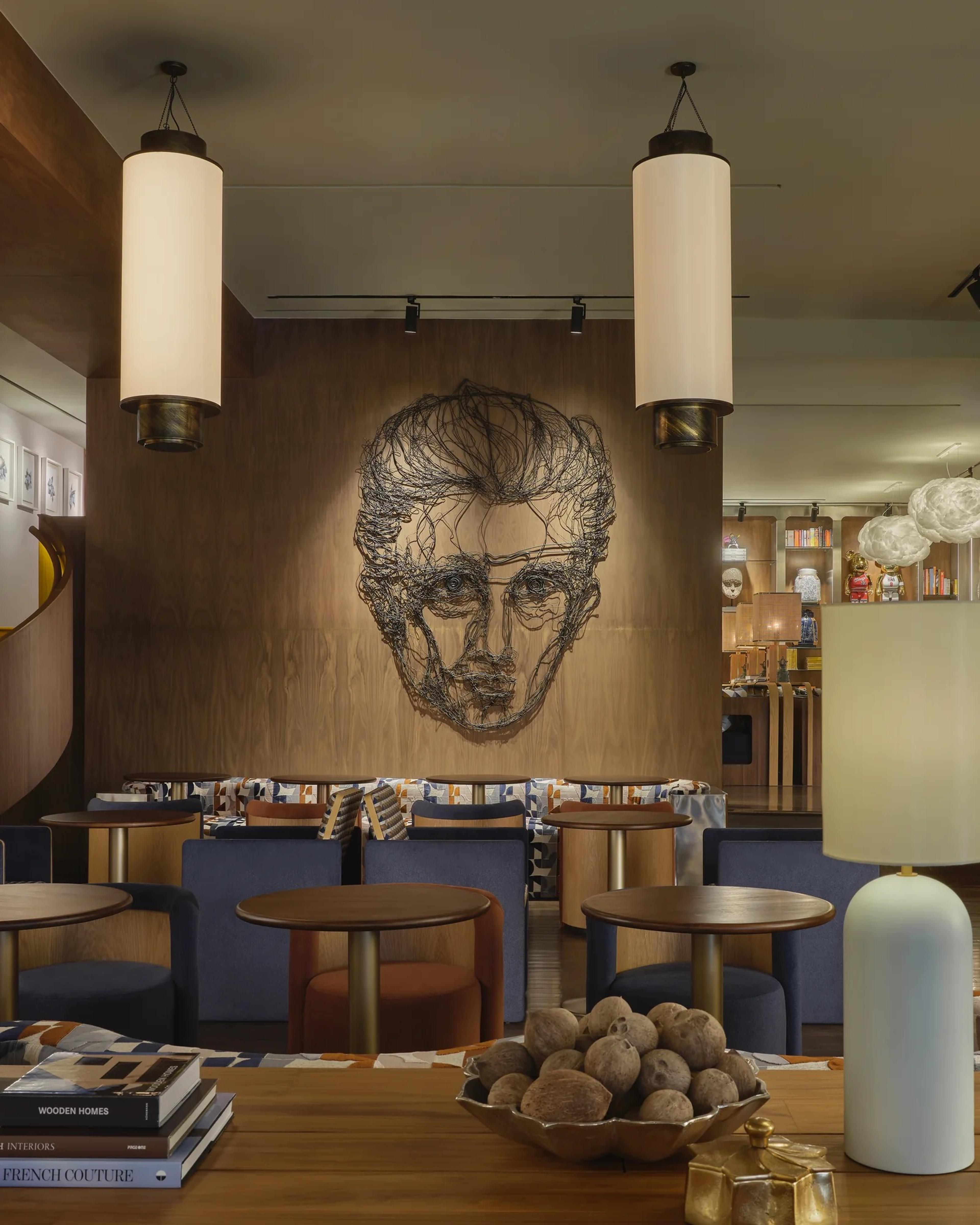 A hotel lounge with cylindrical pendant lights hanging over blue banquette seating, a wire sculpture portrait on the wood panelled wall behind.