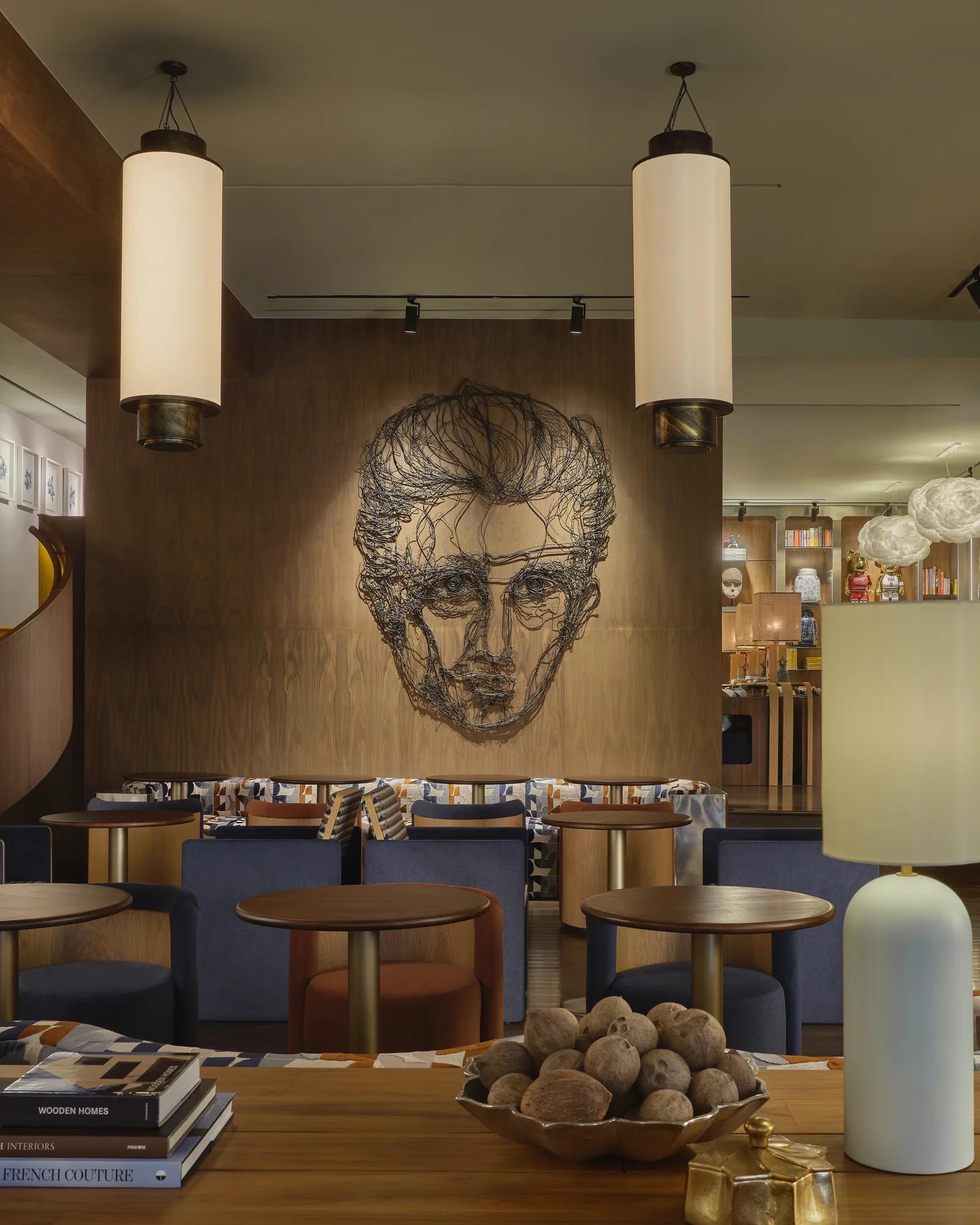 A hotel lounge with cylindrical pendant lights hanging over blue banquette seating, a wire sculpture portrait on the wood panelled wall behind.
