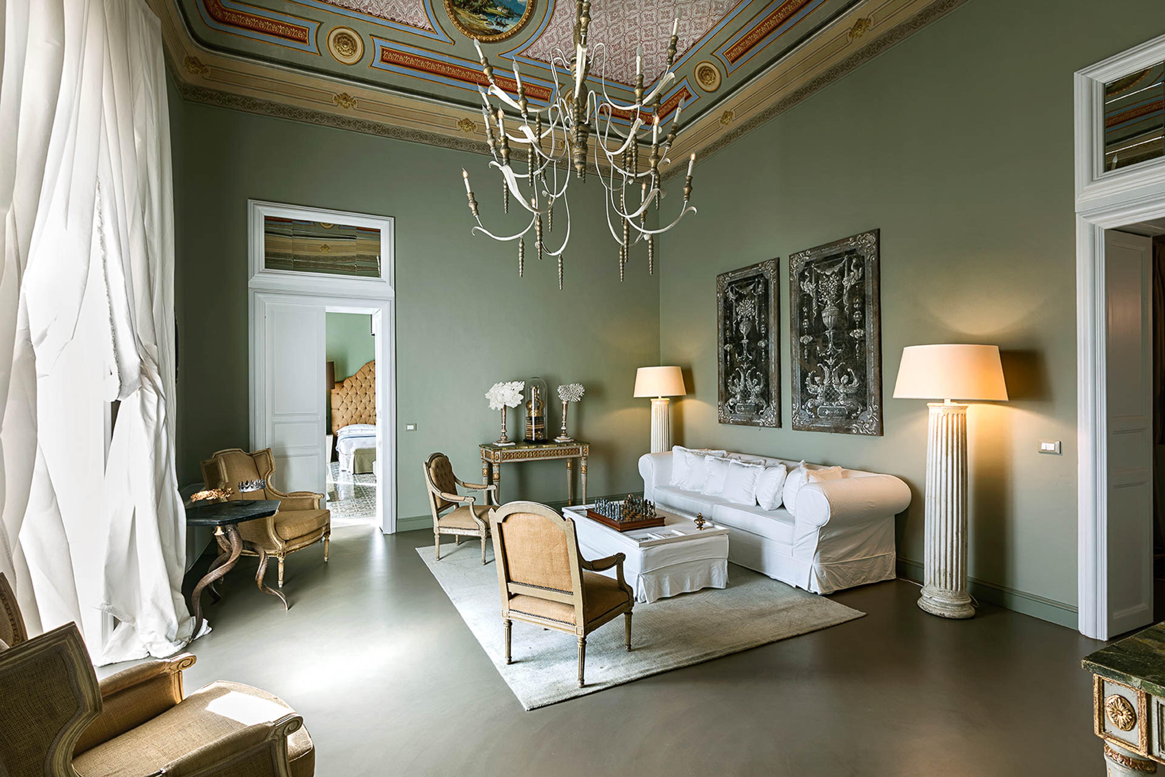 A sitting room with a painted and gilded ceiling, a crystal chandelier, a white slipcovered sofa and armchairs arranged on a patterned rug.