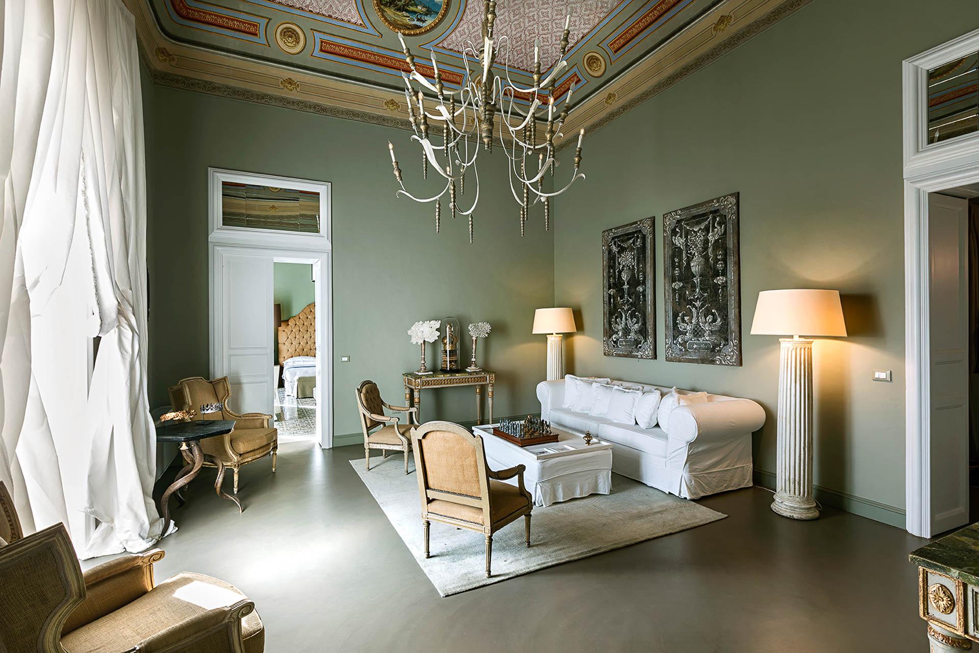 A sitting room with a painted and gilded ceiling, a crystal chandelier, a white slipcovered sofa and armchairs arranged on a patterned rug.