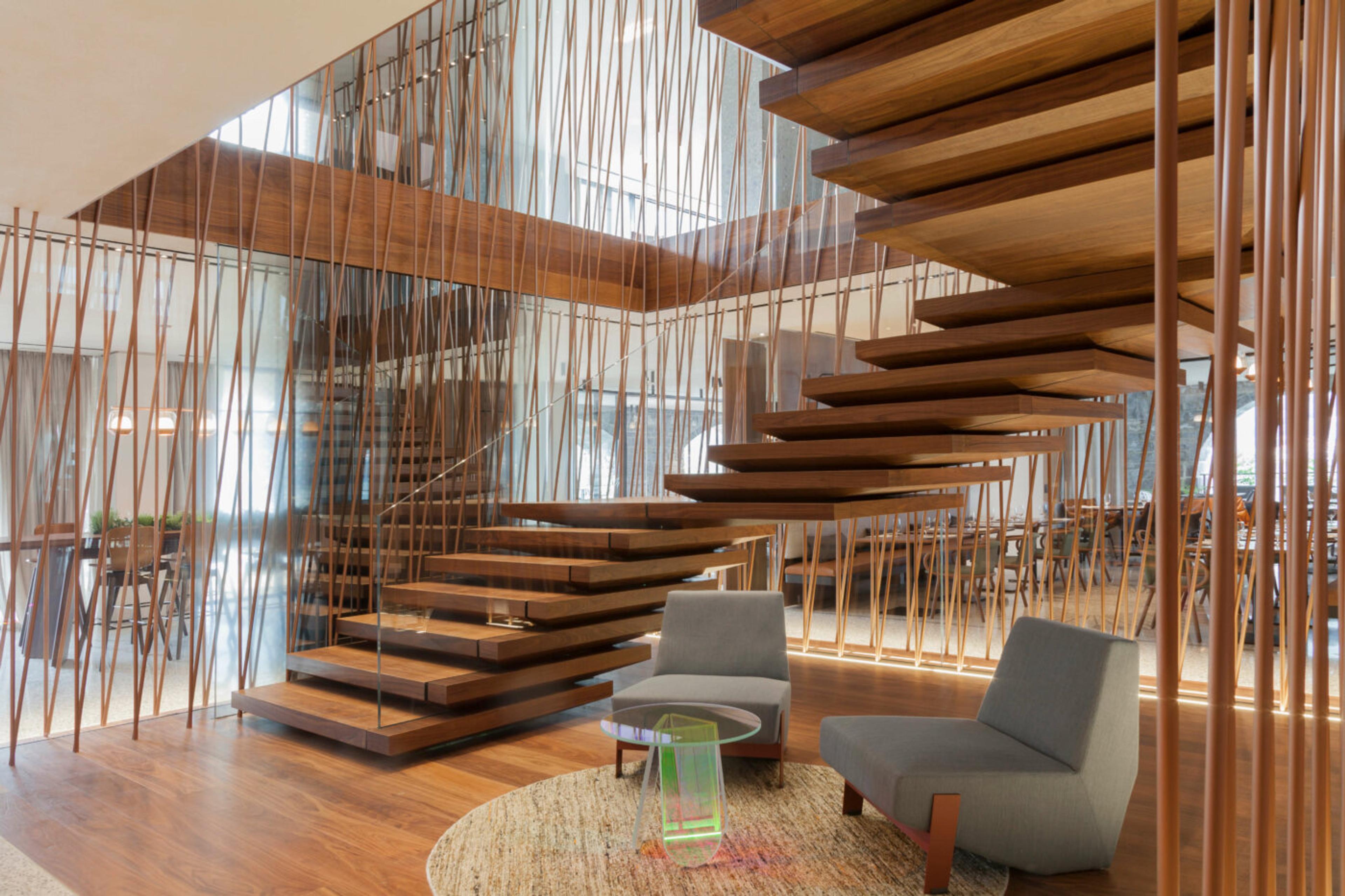 A freestanding timber staircase with floating wooden treads, strung between vertical dowels, in a double-height room with two grey lounge chairs below.