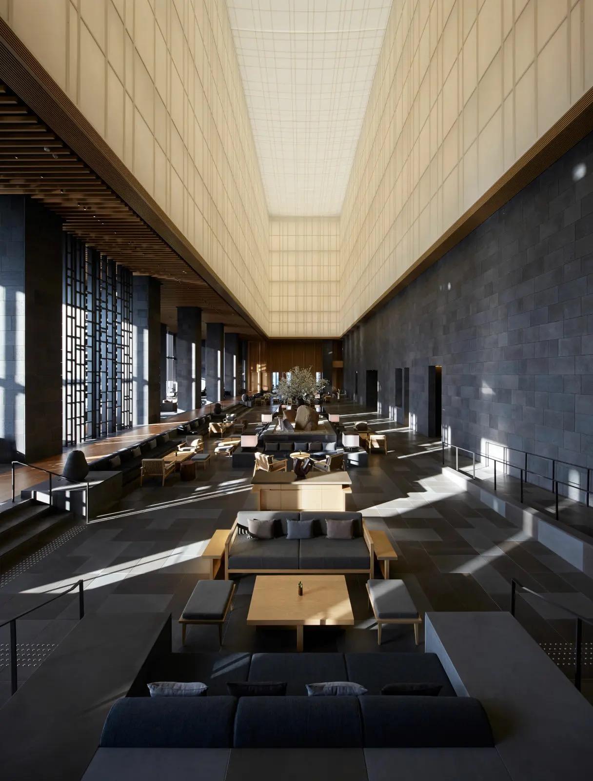 A long, soaring hotel lobby with a glowing fabric ceiling, dark stone walls on one side and floor-to-ceiling screened windows on the other, furnished with low grey sofas and wooden tables.