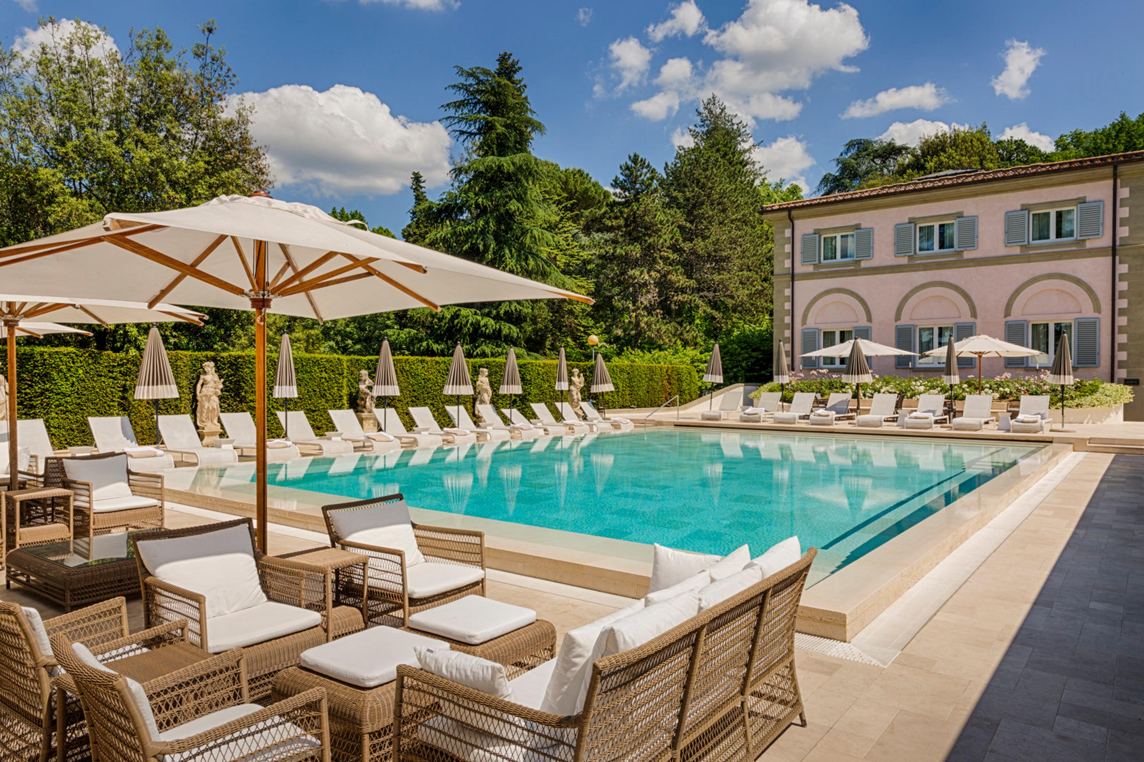 A rectangular pool with white parasols and loungers along its edge, a wicker sofa set in the foreground, and a pale pink building on the right.