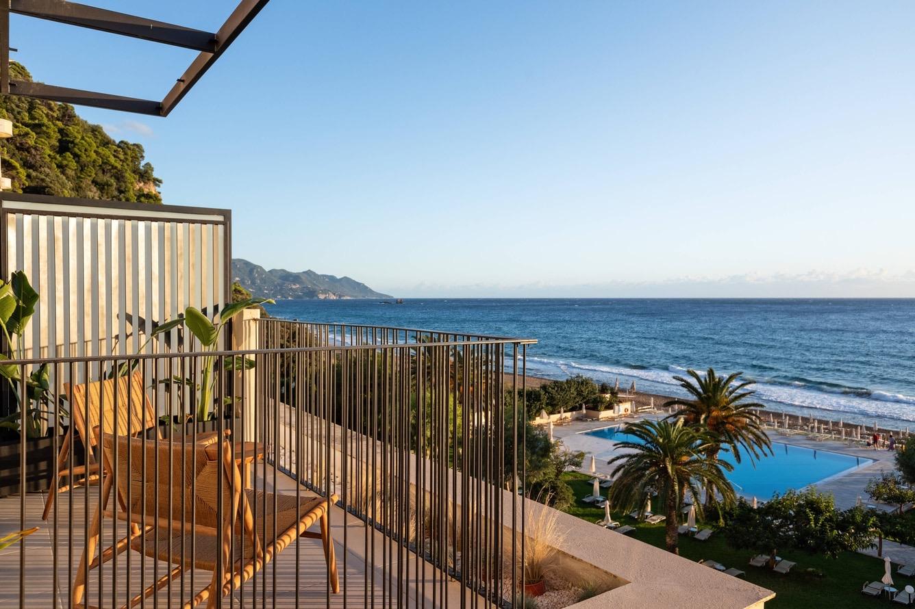 A high balcony view over a wooden pergola and potted plants to a pool, gardens, a pine-covered headland, and the sea beyond.