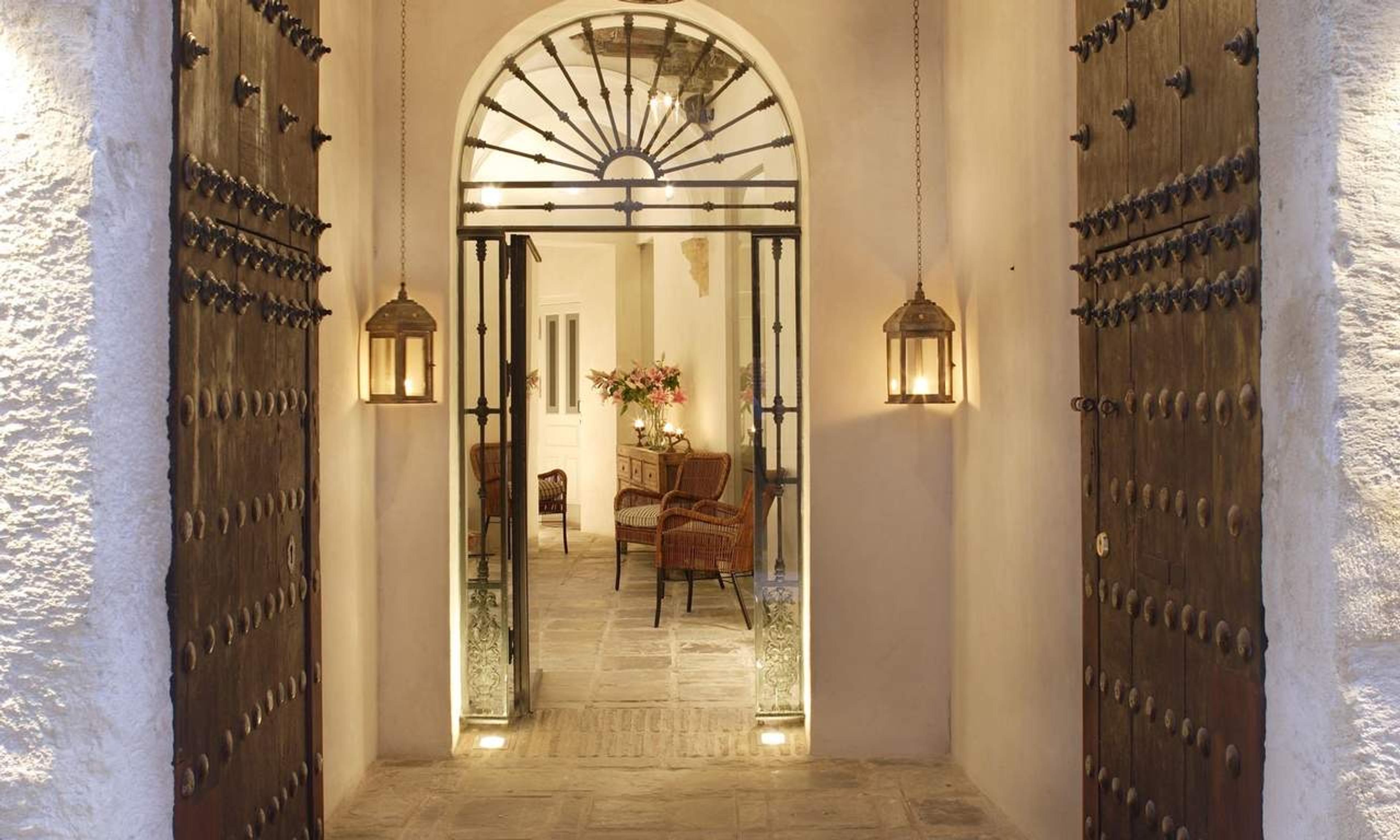 An arched doorway with studded wooden doors open to a tiled hallway lit by hanging lanterns, with wicker chairs and a vase of pink flowers beyond.
