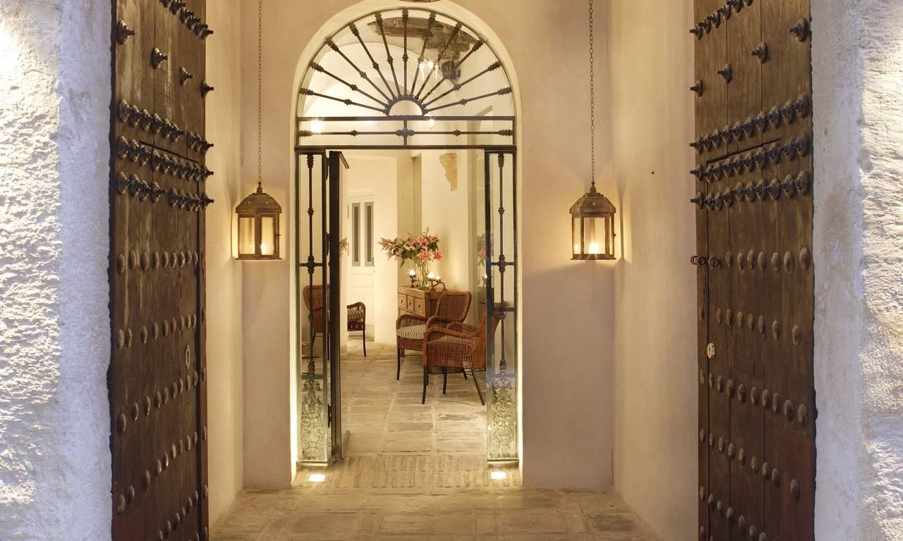 An arched doorway with studded wooden doors open to a tiled hallway lit by hanging lanterns, with wicker chairs and a vase of pink flowers beyond.