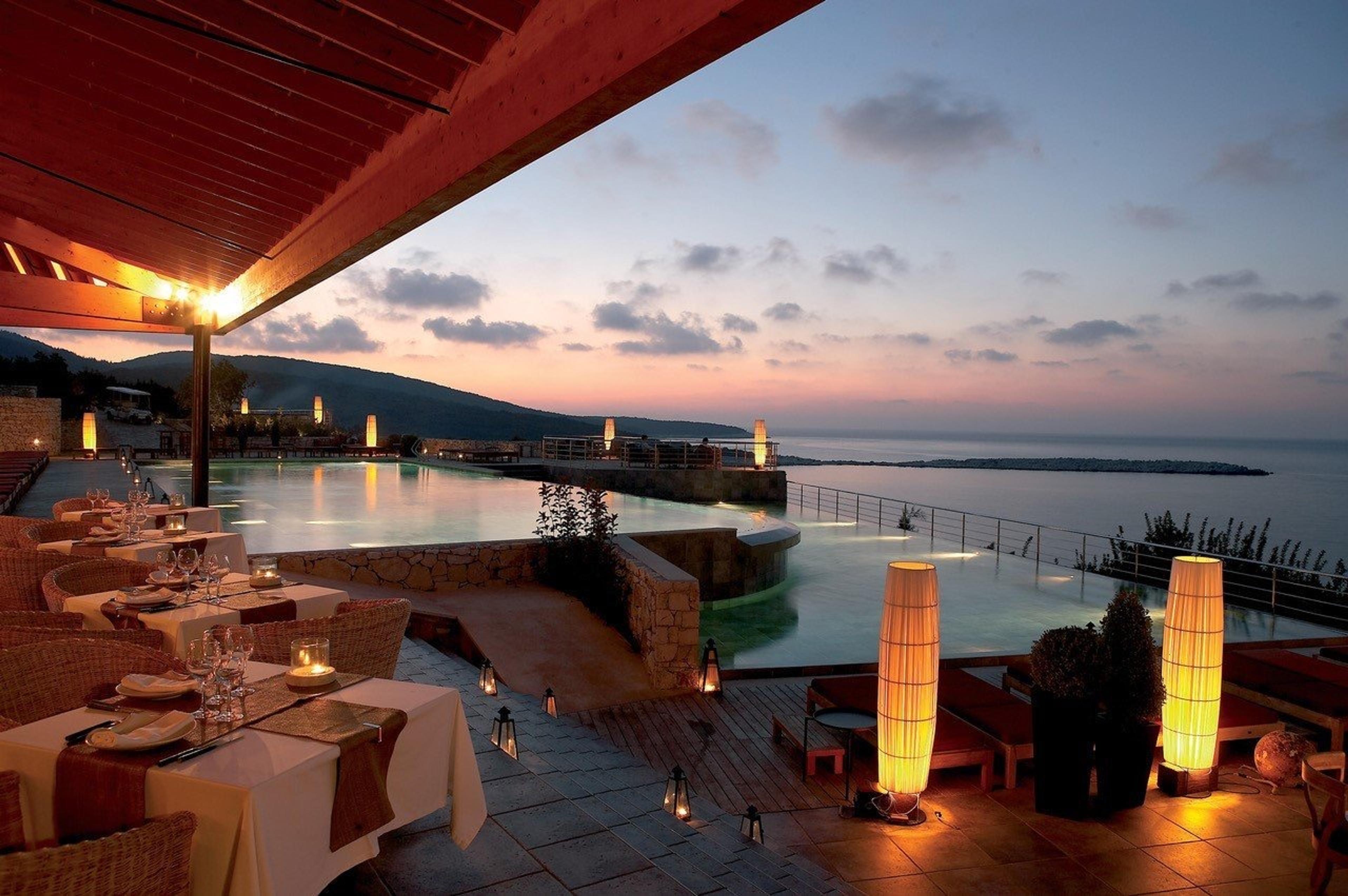 A restaurant terrace at dusk, tables laid with white cloths and glassware beside an infinity pool and the sea.