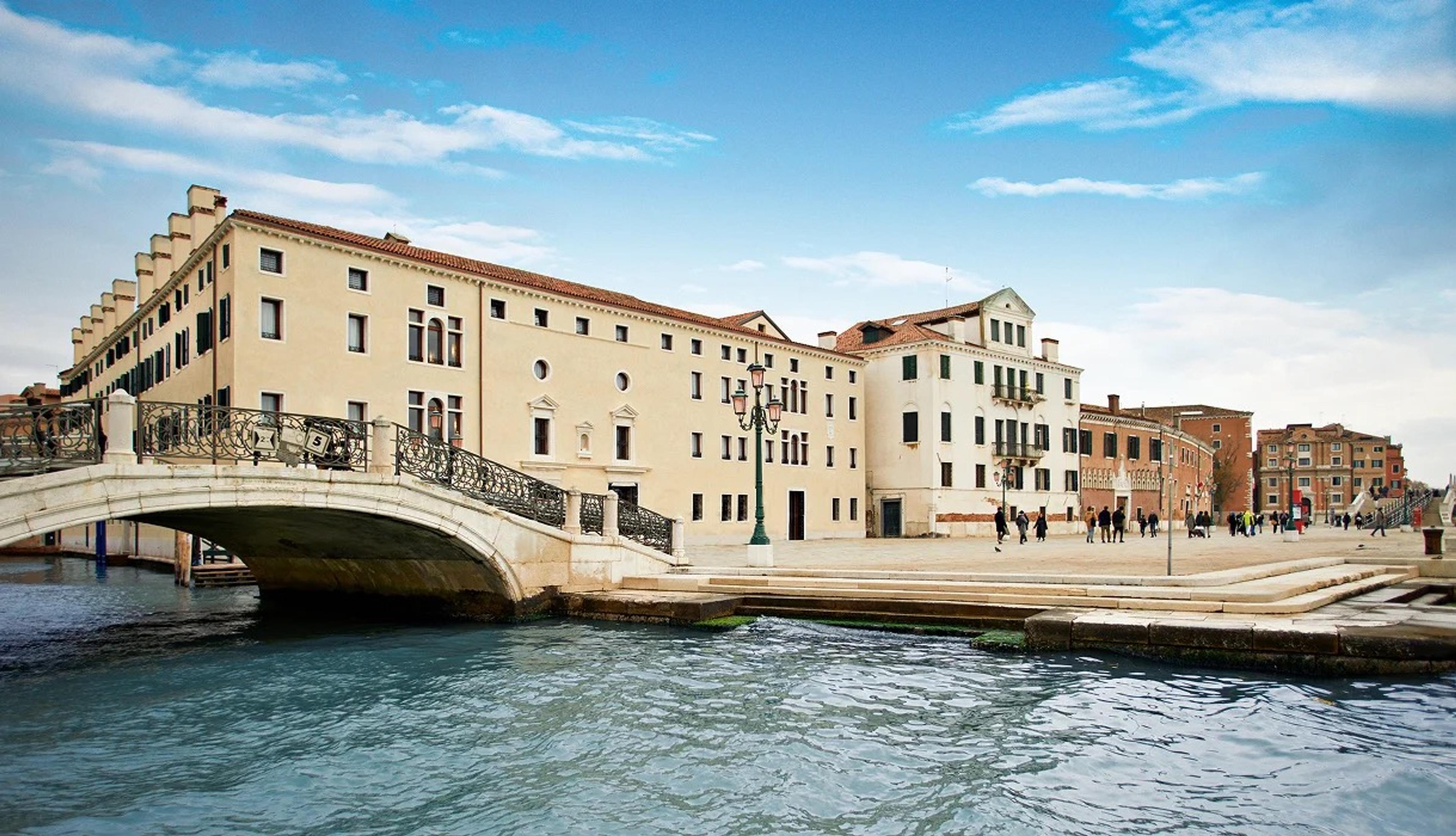 A stone bridge over a canal leading to a long pale building with arched windows, people walking along the waterfront.