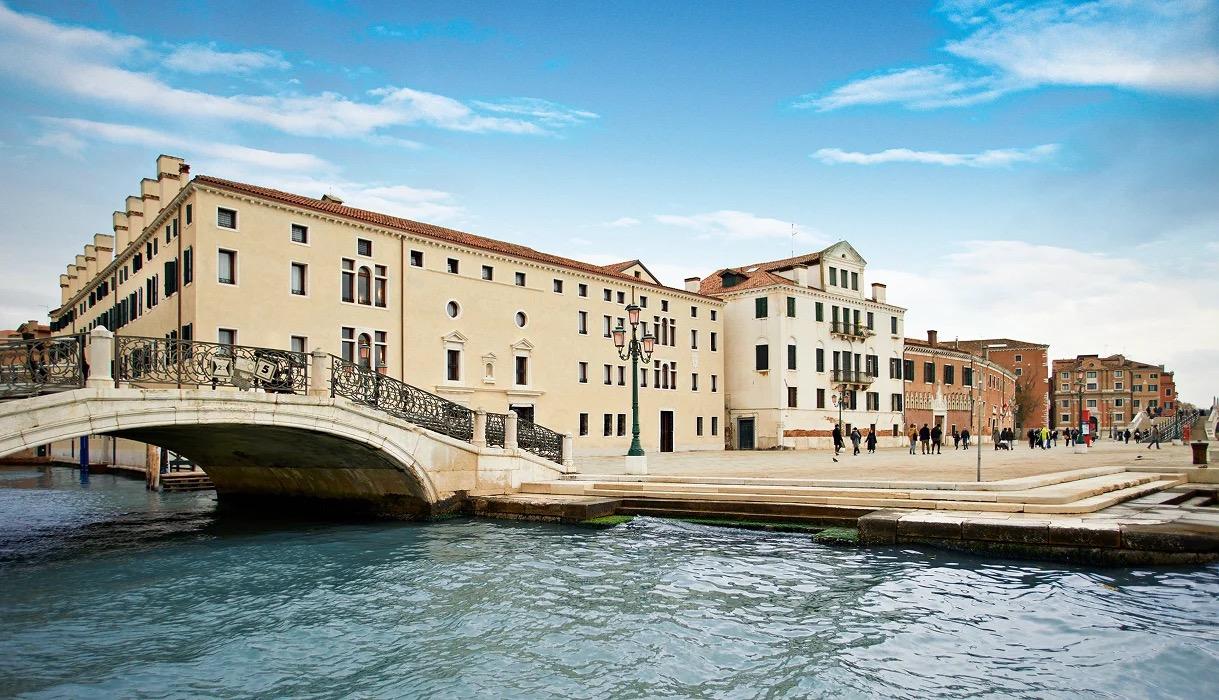 A stone bridge over a canal leading to a long pale building with arched windows, people walking along the waterfront.