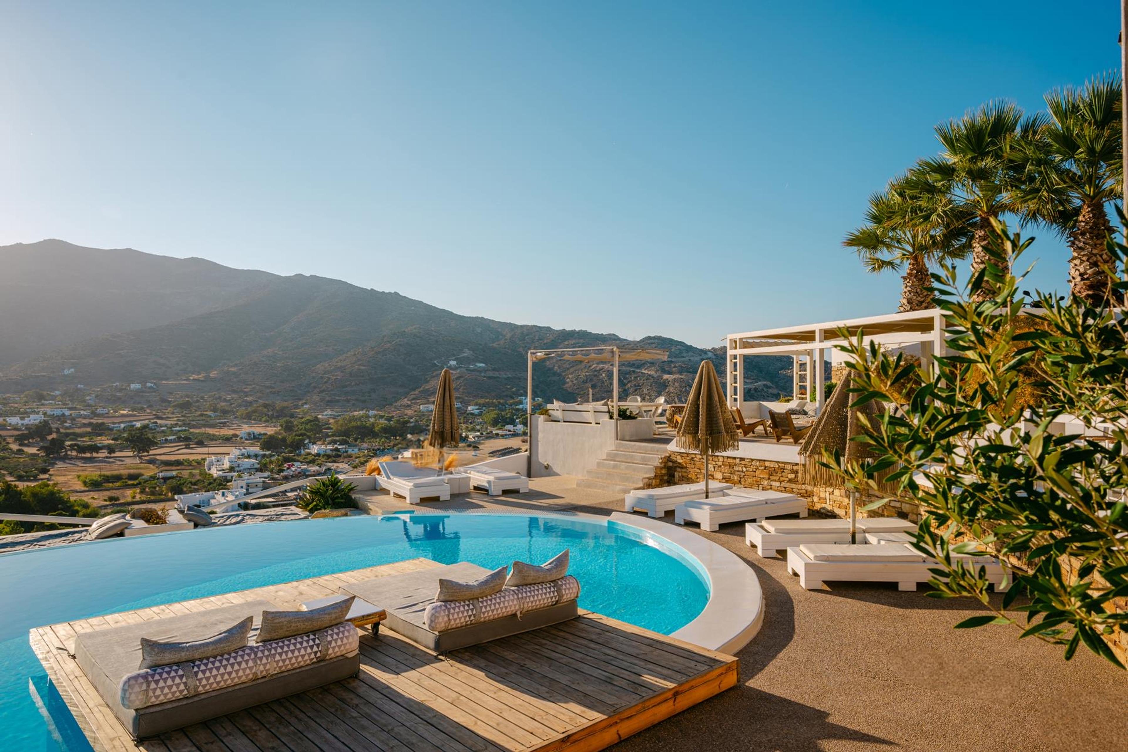 An infinity pool on a timber deck with two cushioned loungers, palm trees and a pergola on the terrace, and a dry mountain behind the village in the valley below.