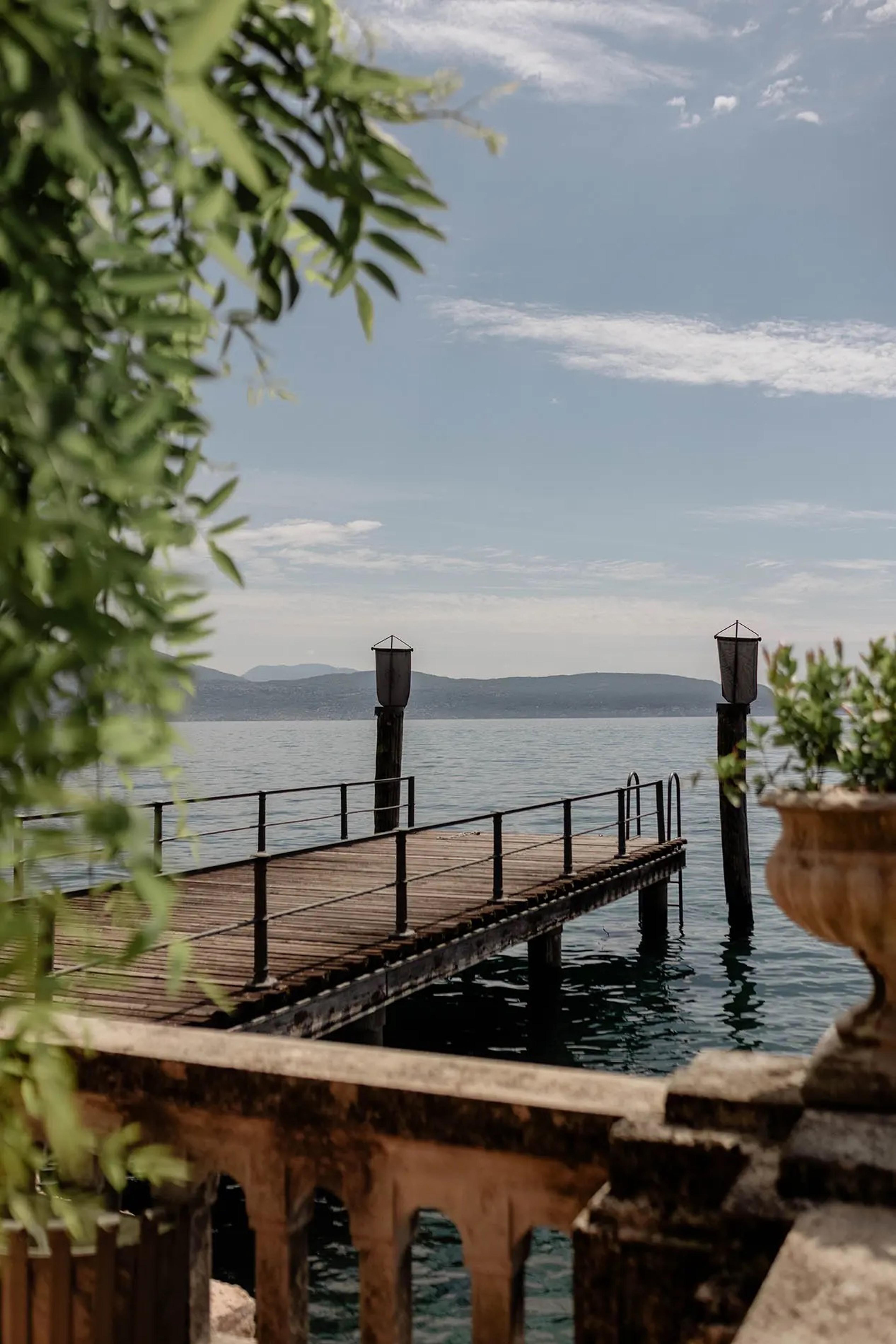 A wooden pier with two lamp posts extends from a stone balustrade into open lake water, hills visible on the far shore.