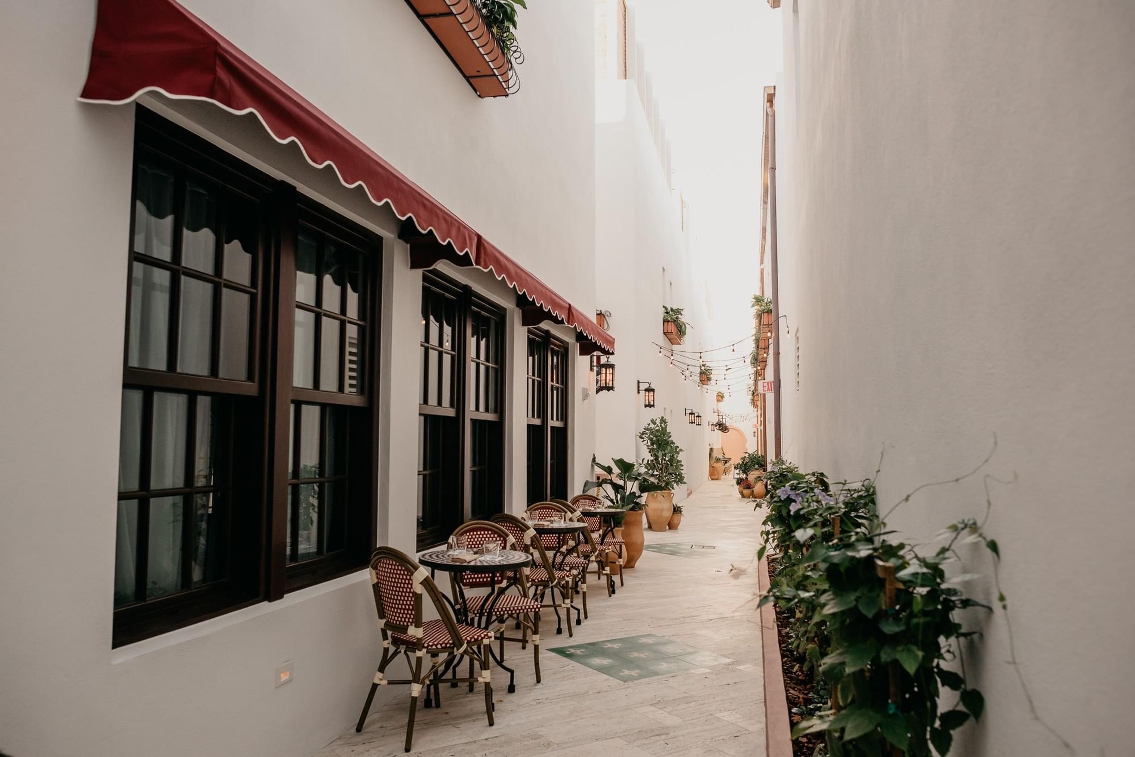 A narrow courtyard with a red awning, cafe tables and chairs, potted plants, and string lights strung between the walls.