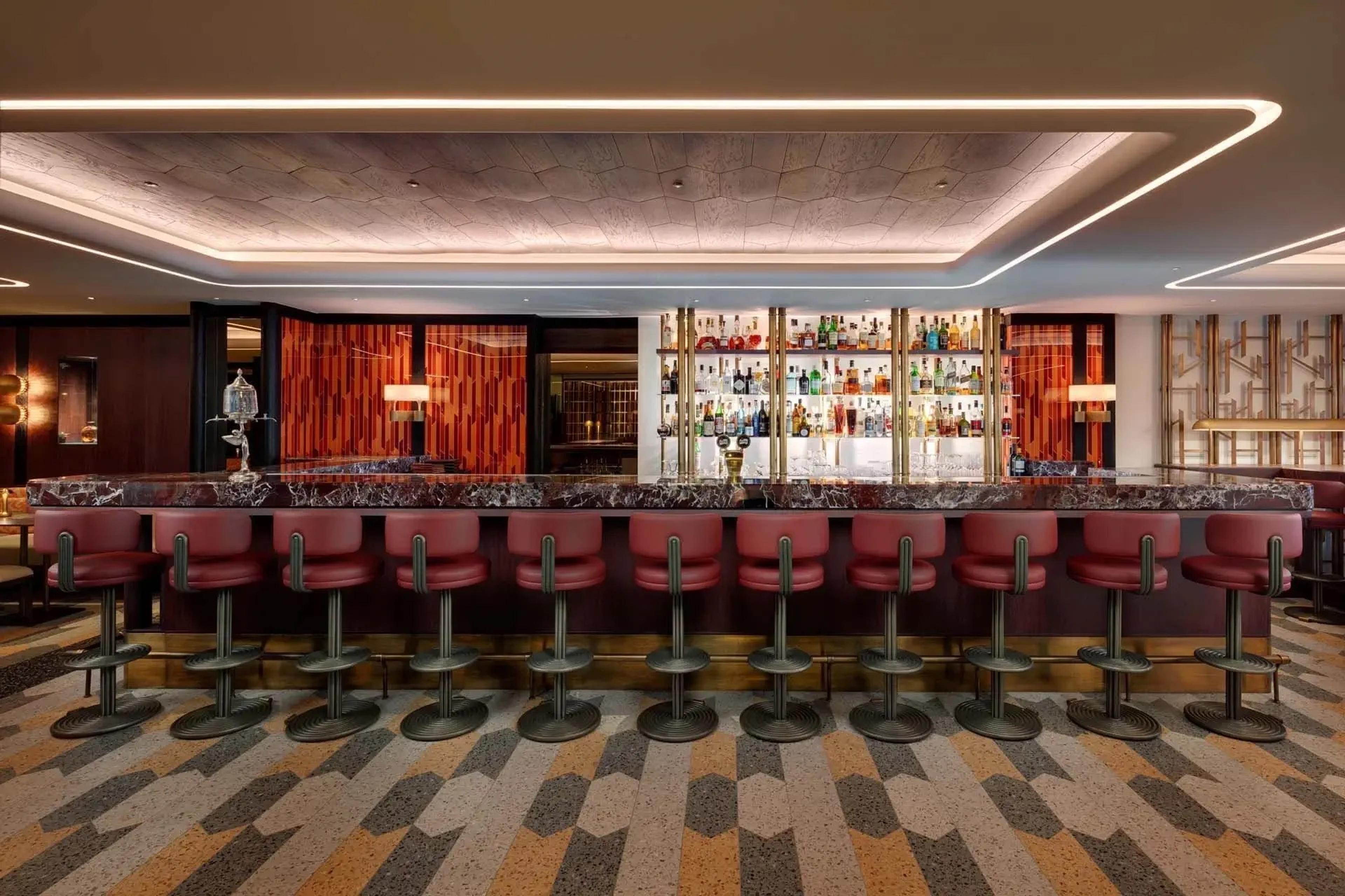 A bar with a long marble counter, a row of red leather stools on a patterned carpet, and bottles on a backlit shelf.
