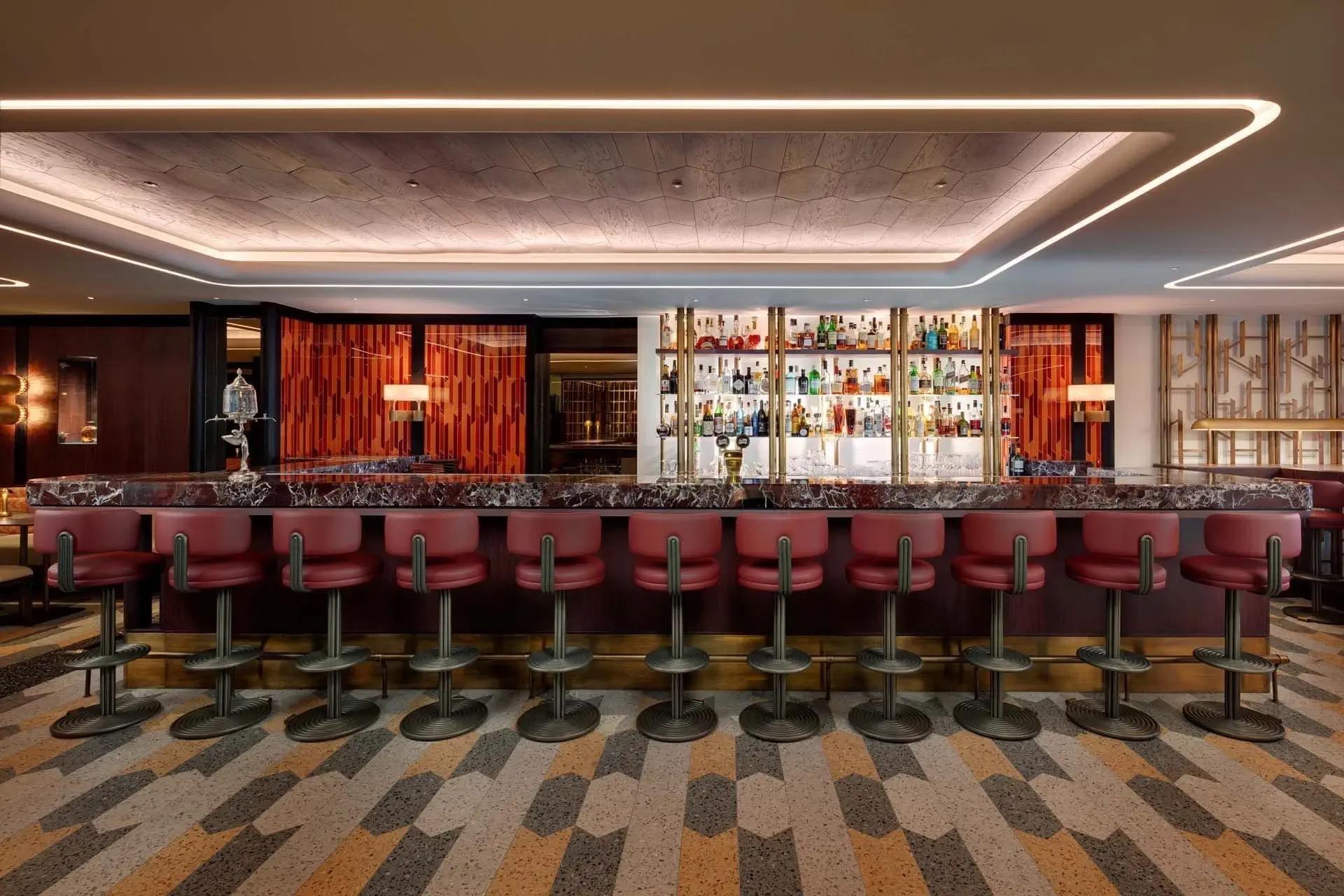 A bar with a long marble counter, a row of red leather stools on a patterned carpet, and bottles on a backlit shelf.