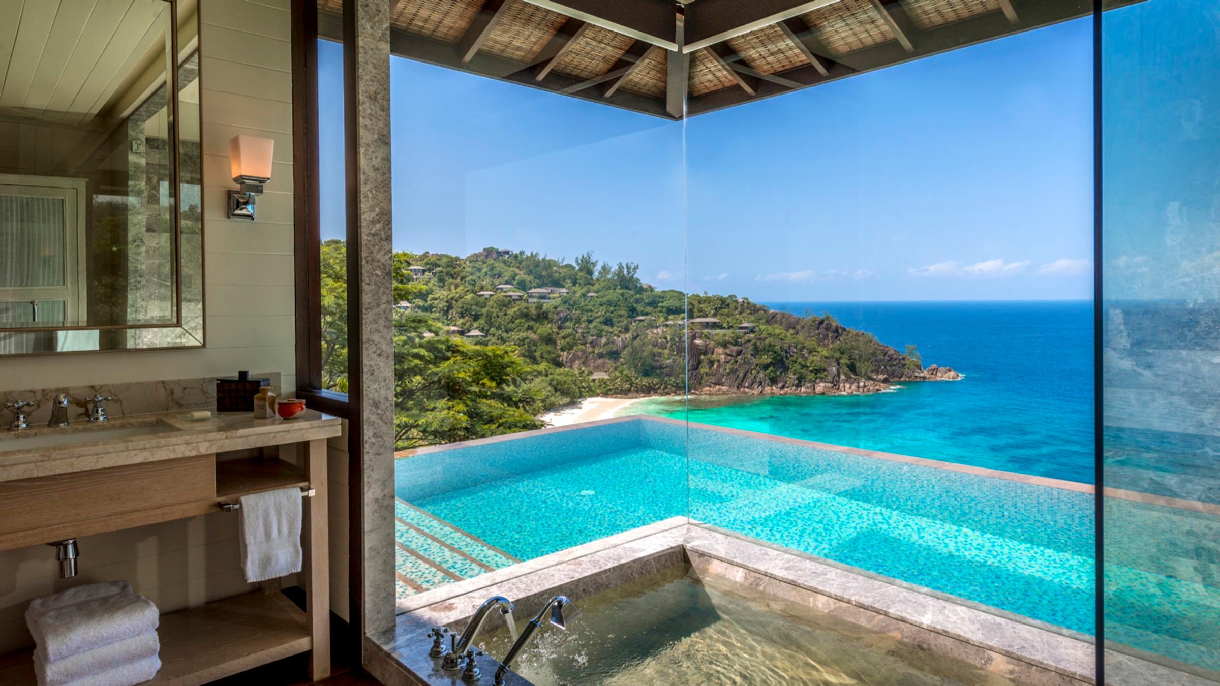 A bathroom with a stone tub set beside a glass wall, so the water appears to merge with an infinity pool and the sea beyond.