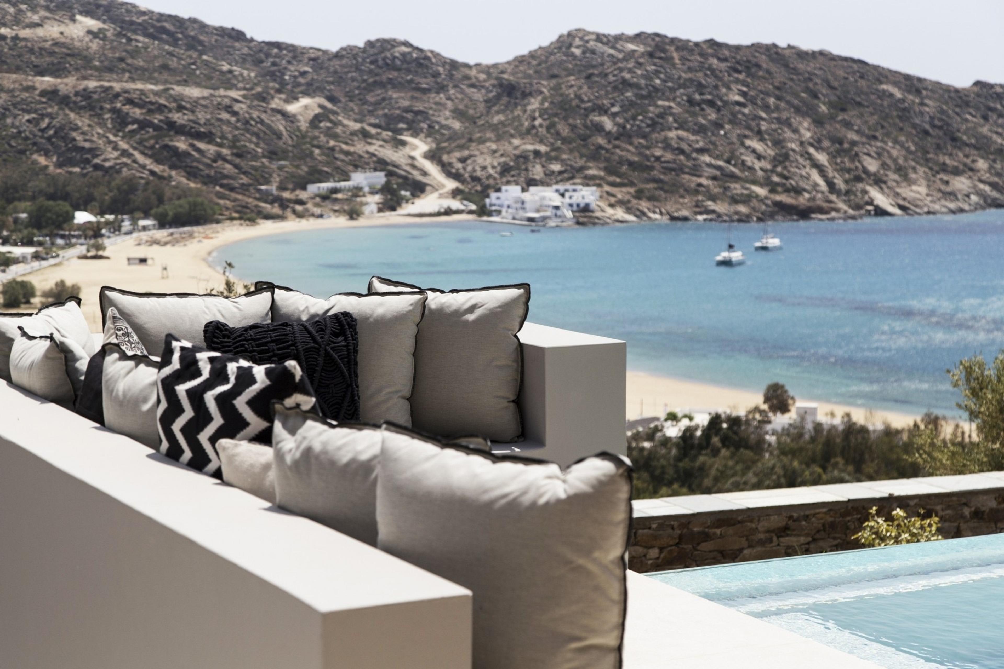 Cushions on a white ledge in the foreground, with a sandy bay, boats on blue water and white houses on the shore beyond.