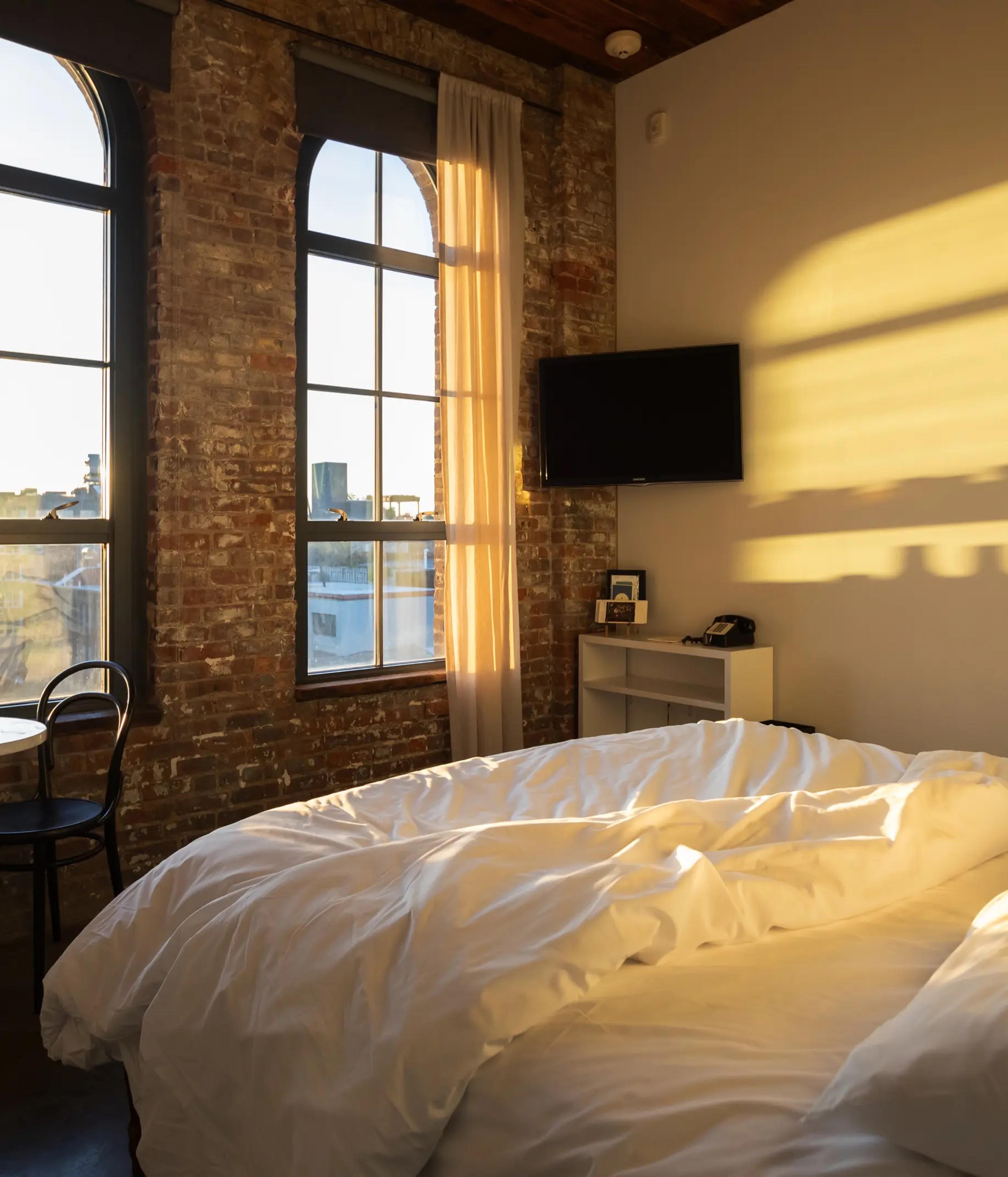 An unmade white bed beside arched brick framed windows, morning light casting long shadows across the wall and a wall mounted television.