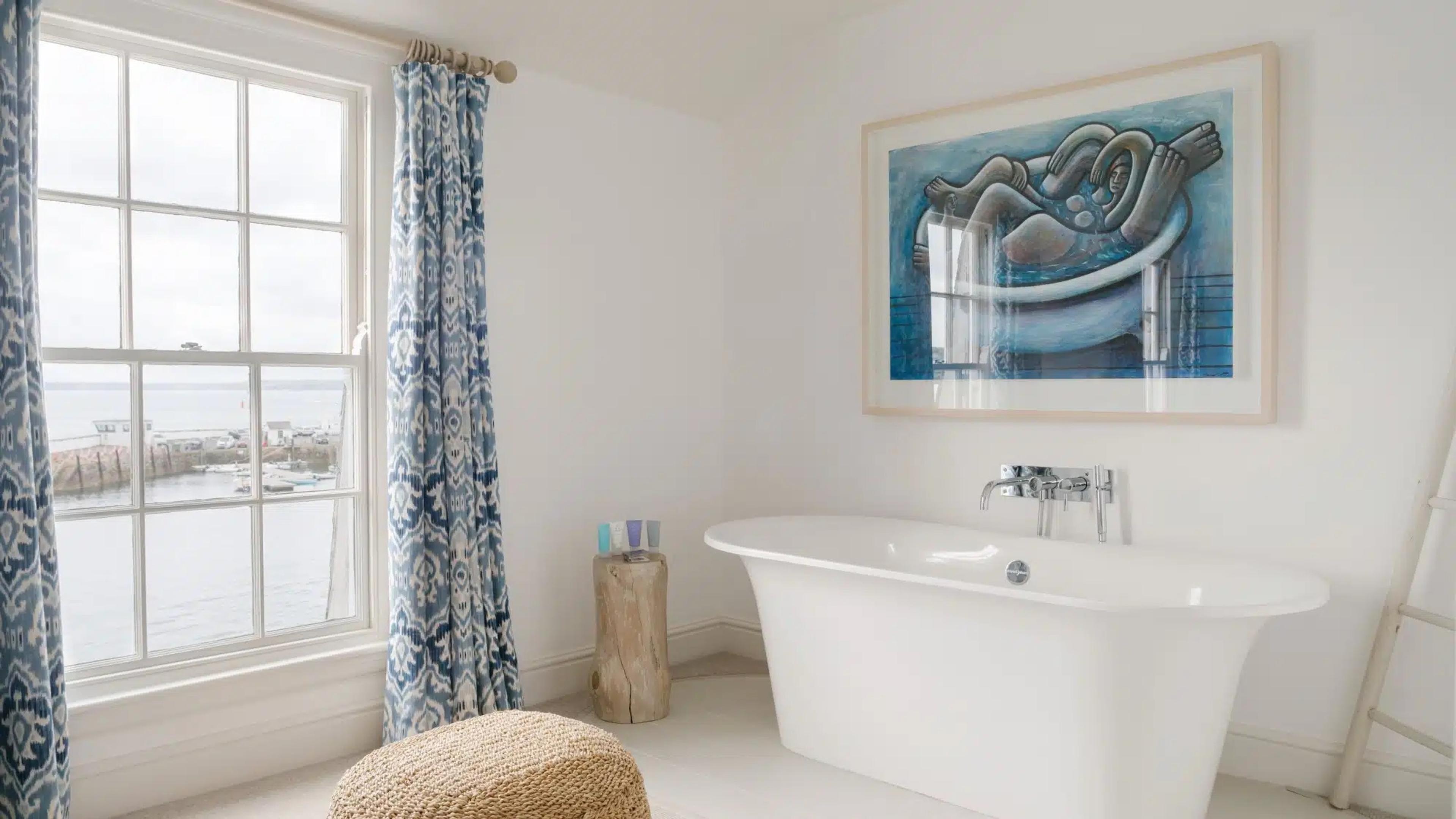 A free-standing white bathtub below a window looking out over a harbour full of moored boats, with patterned blue curtains and a framed painting on the wall.